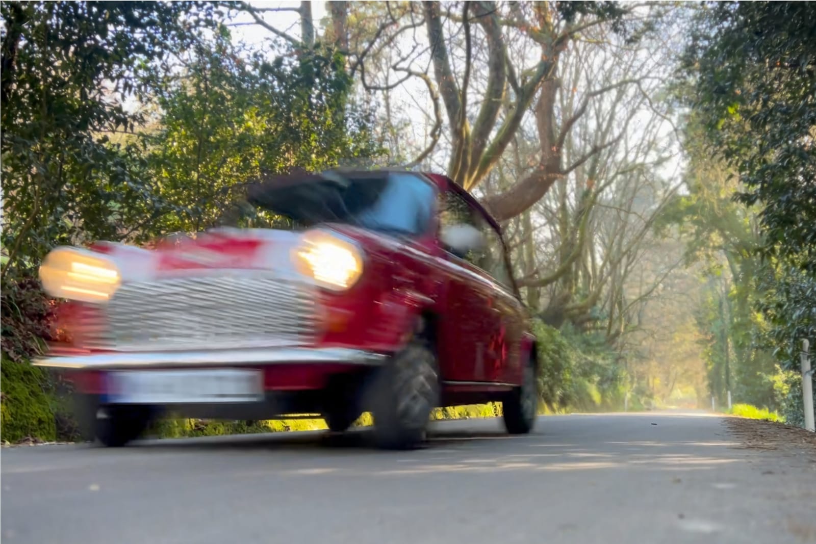 Red Classic Mini at speed on a country road, headlights on, the trees blurred with motion.