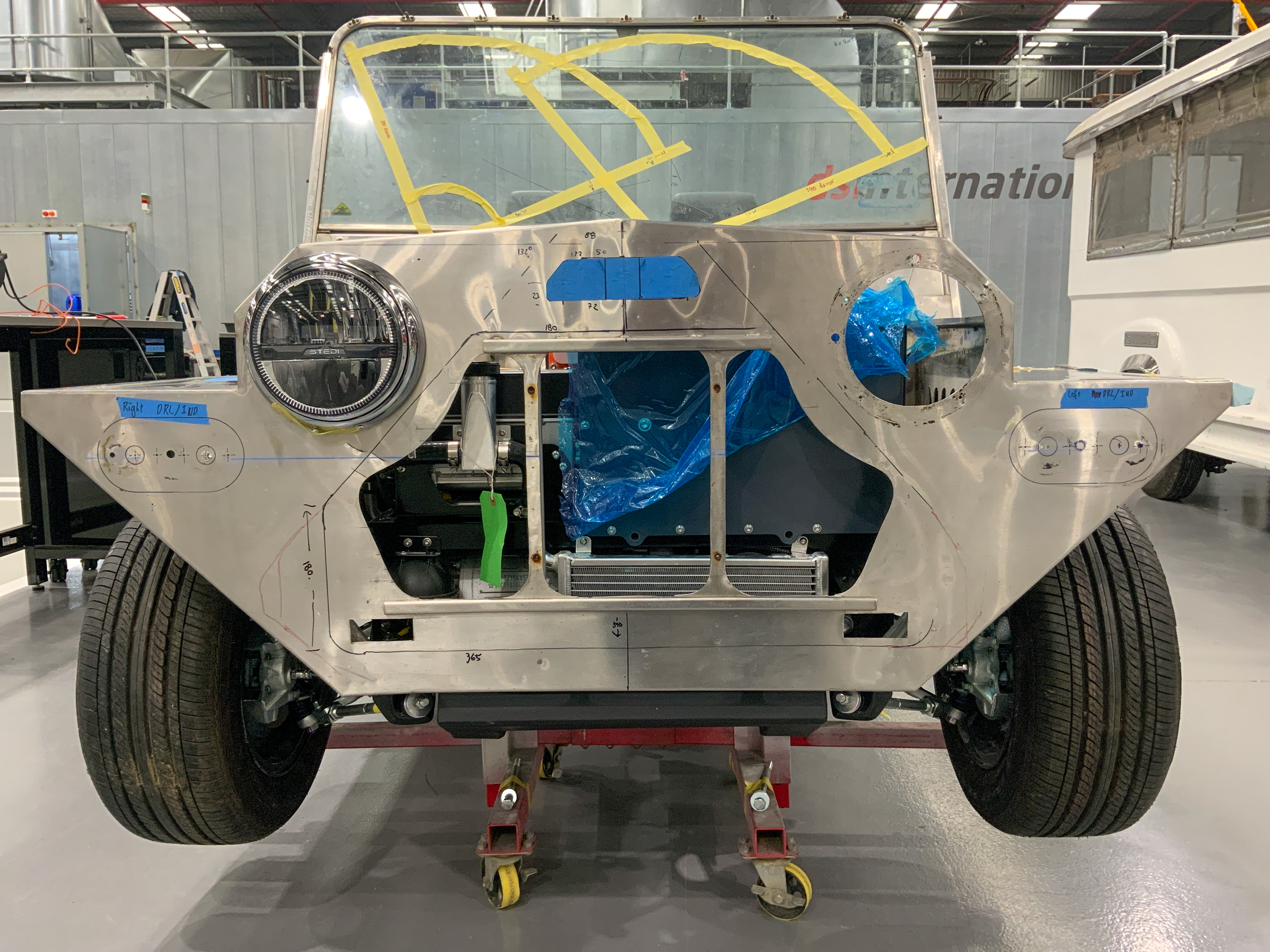 Front-on view of the bare stainless body shell with the battery bay open and protected by blue tape, masked windscreen frame, and builder's layout markings visible.