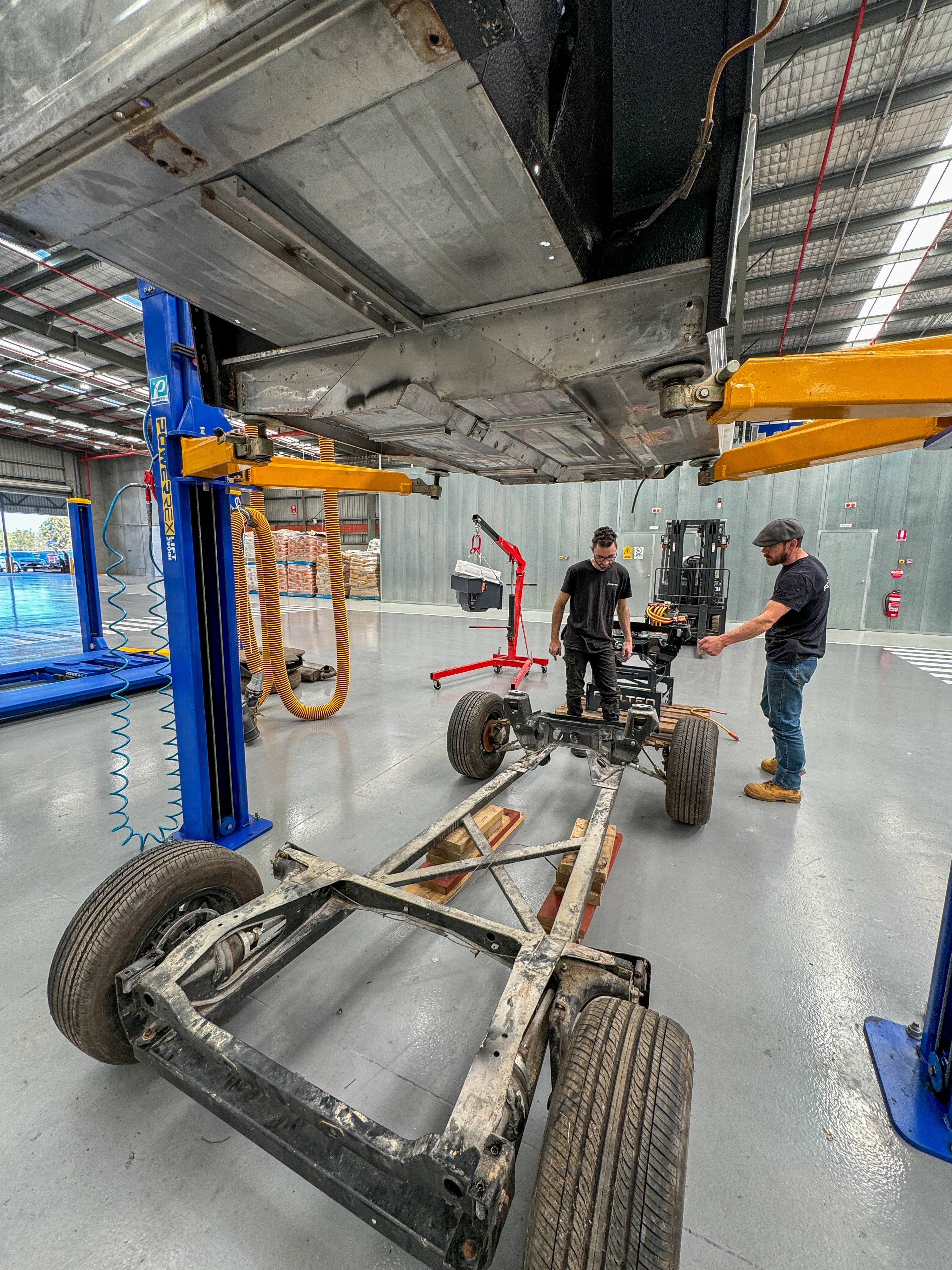 Two technicians work on the bare rolling chassis after the body has been removed, with the subframe on the workshop floor and a hoist gantry overhead.