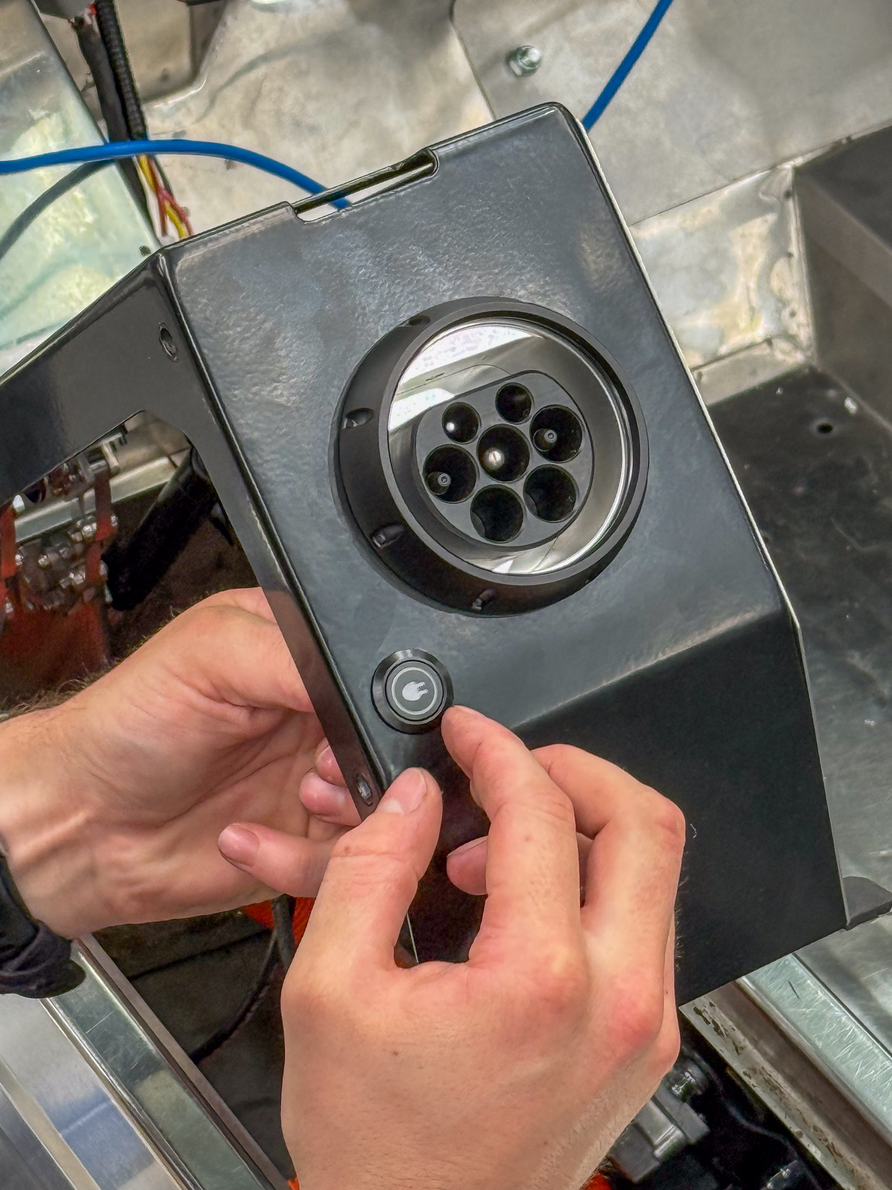 Technician's hands fitting the CCS2 charge port module into the side-pod housing during assembly, with blue wiring and the stainless body panel surrounding the work area.