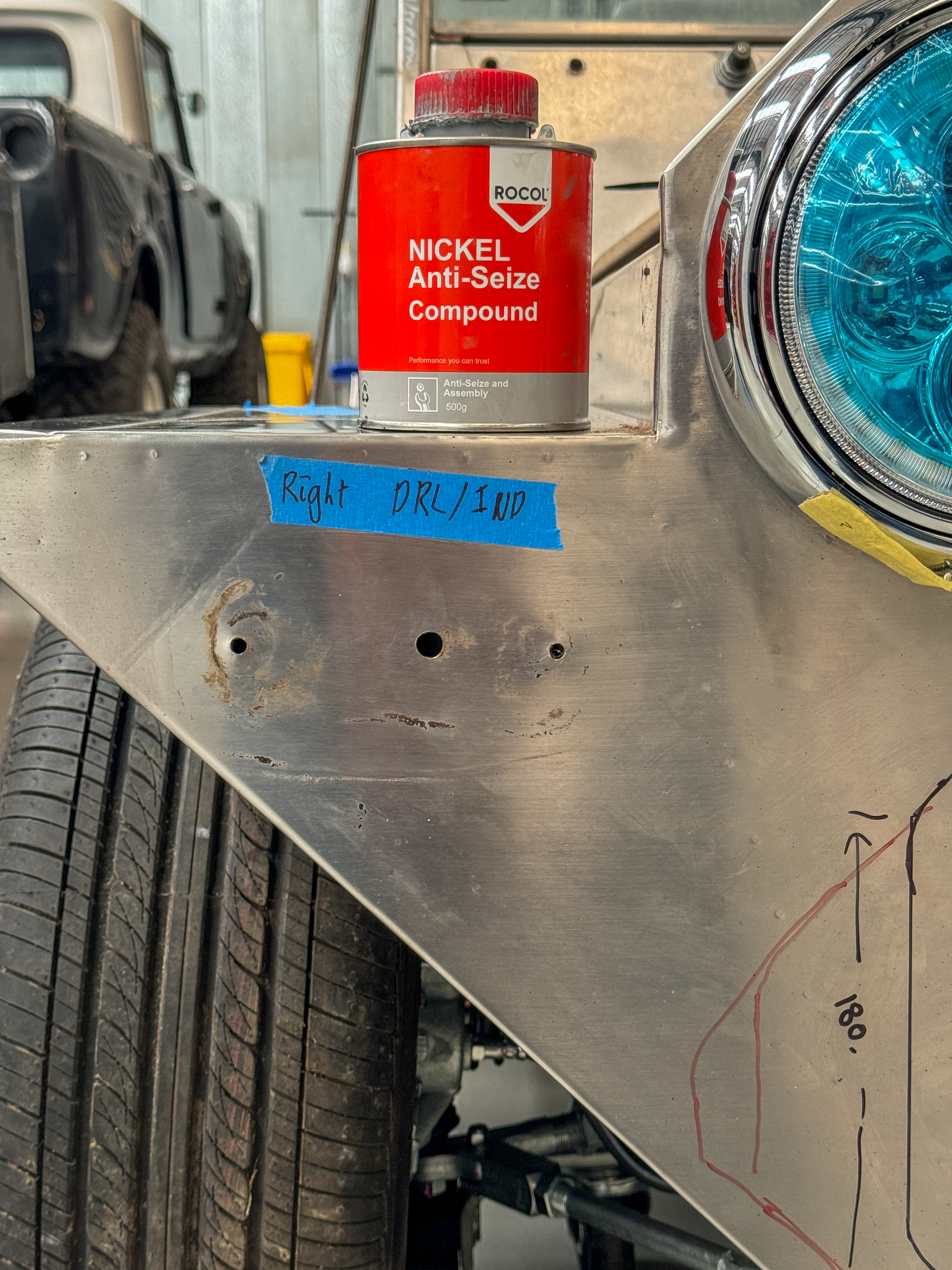 Early-build front panel with DRL and indicator positions marked in pen on masking tape, a teal-lensed headlight fitted and a can of anti-seize compound resting on the guard.
