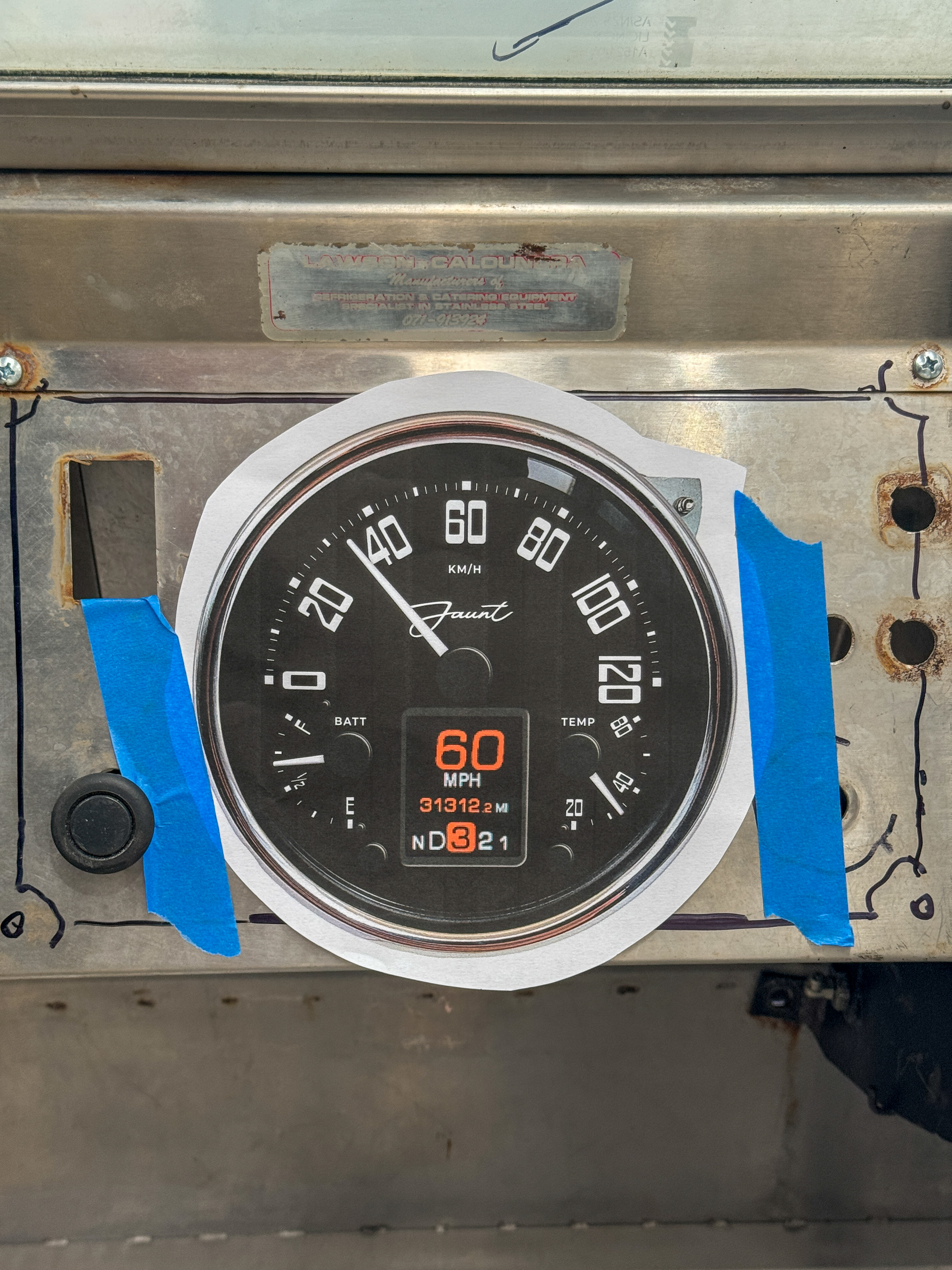 The Jaunt-branded custom speedometer face taped onto the original stainless dashboard panel during design proofing, with the original builder's plate still riveted above.