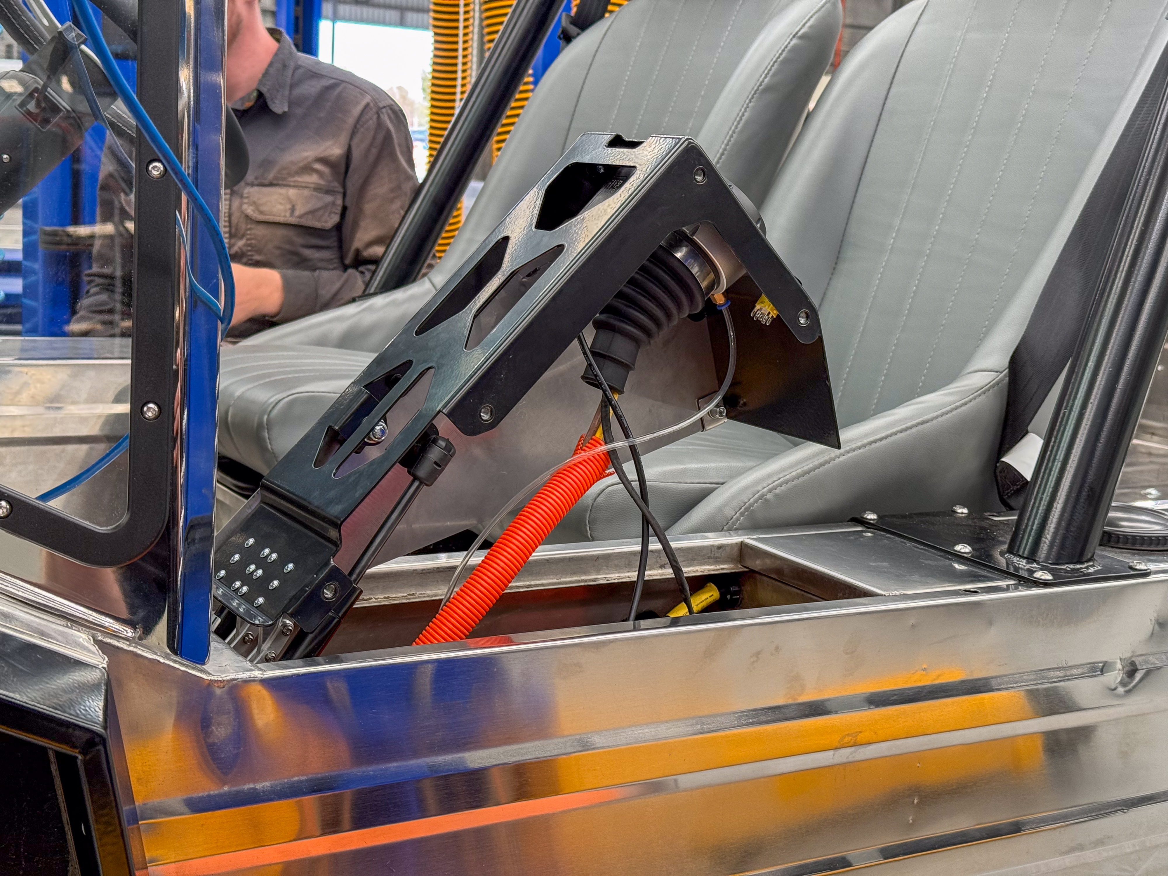 Mid-build interior showing the motor controller bracket bolted between the seats with orange and black wiring partway through installation, hoist arms visible around the body.