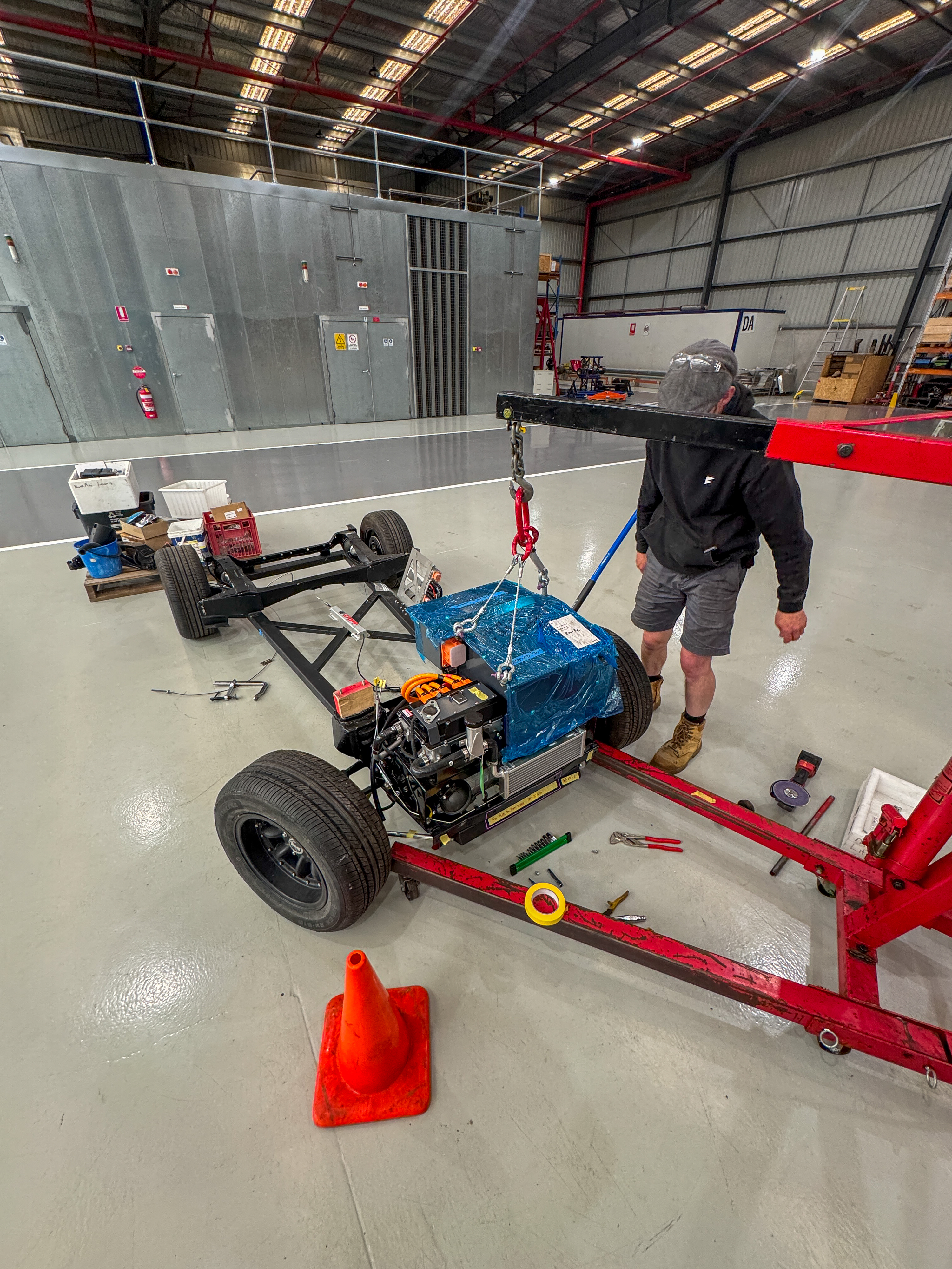 Technician operating a chain hoist to lower the electric motor unit onto the bare rolling chassis in the Jaunt workshop, with a traffic cone and tools on the floor.