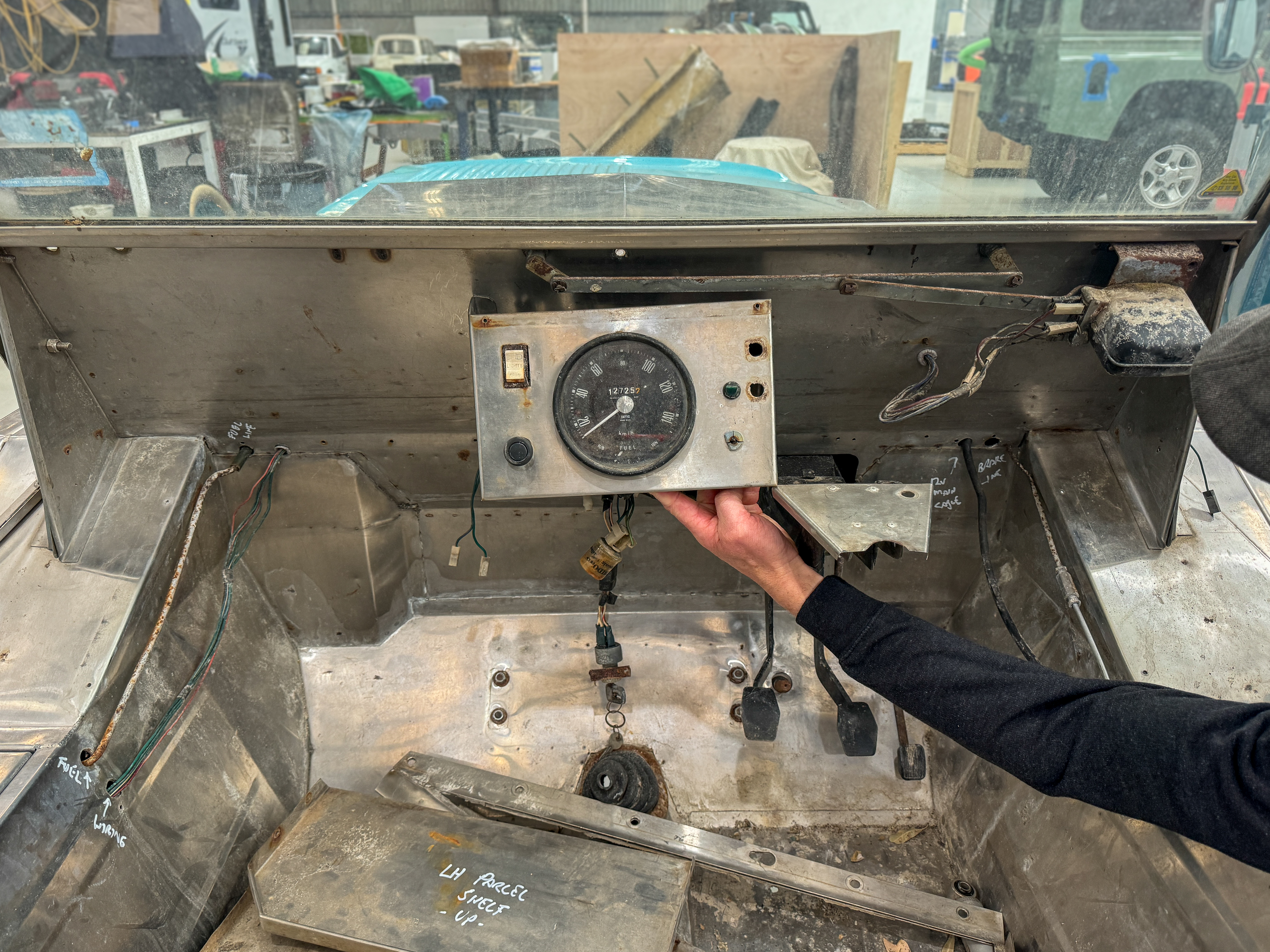 Hand holding the original analogue speedometer cluster against the stripped bare dashboard during the early disassembly phase at the Jaunt workshop.