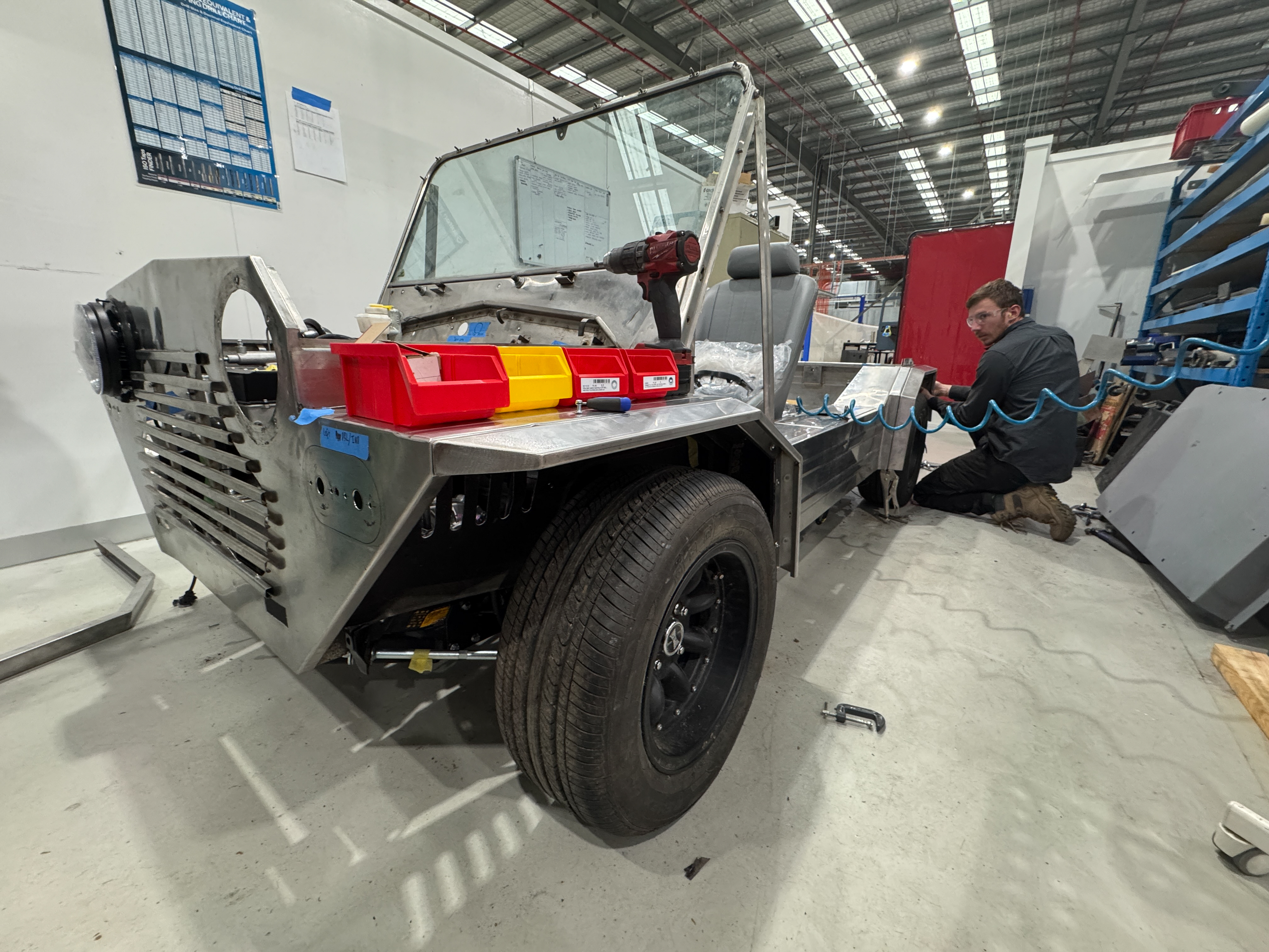 Rear view of the Moke in partial assembly in a smaller workshop, rear taillights taped in position and a technician working at the rear wheel arch.