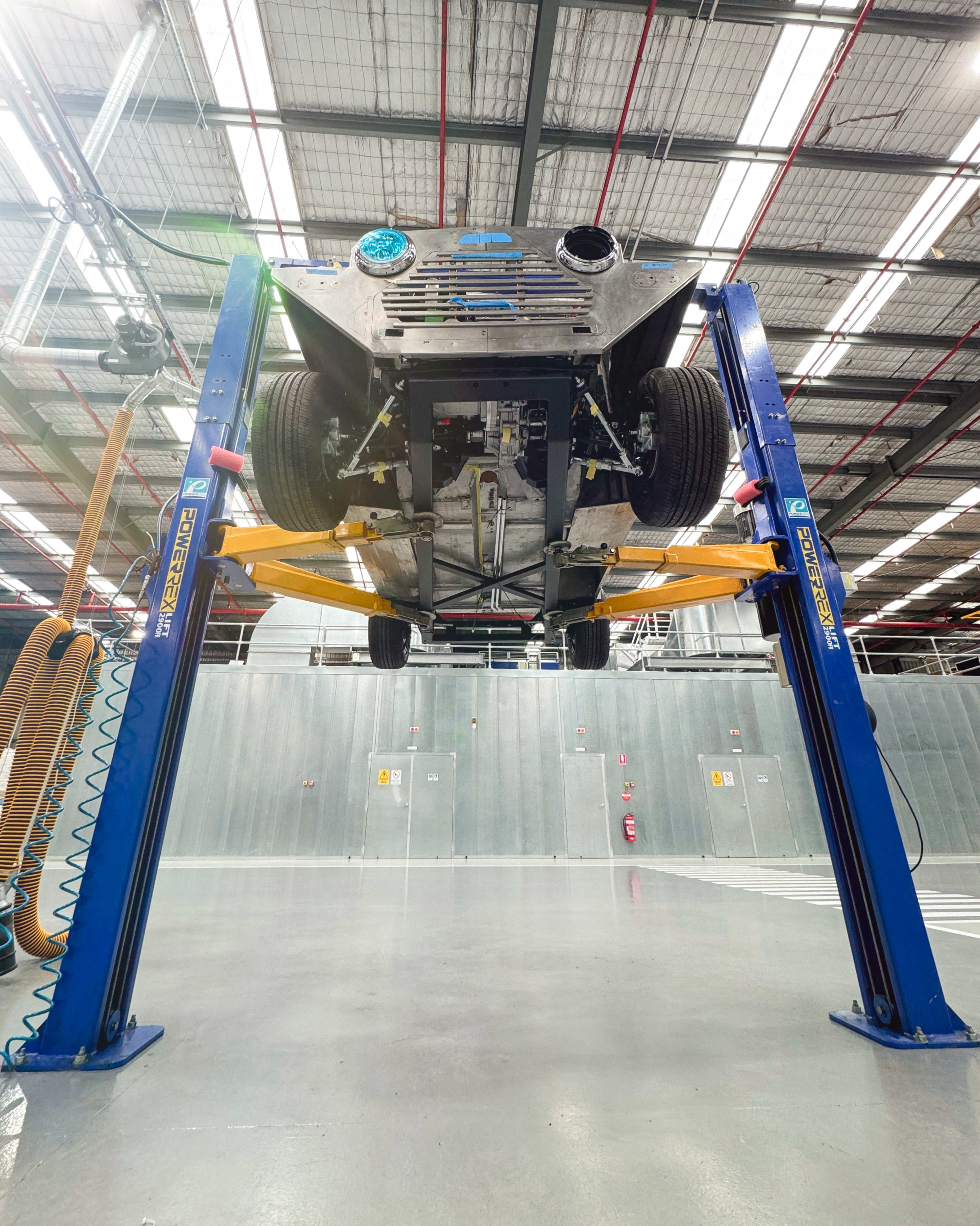 Wide view of the Moke raised on a four-post hoist in the Jaunt workshop, showing the complete underside with battery tray, suspension, and drivetrain from floor level.