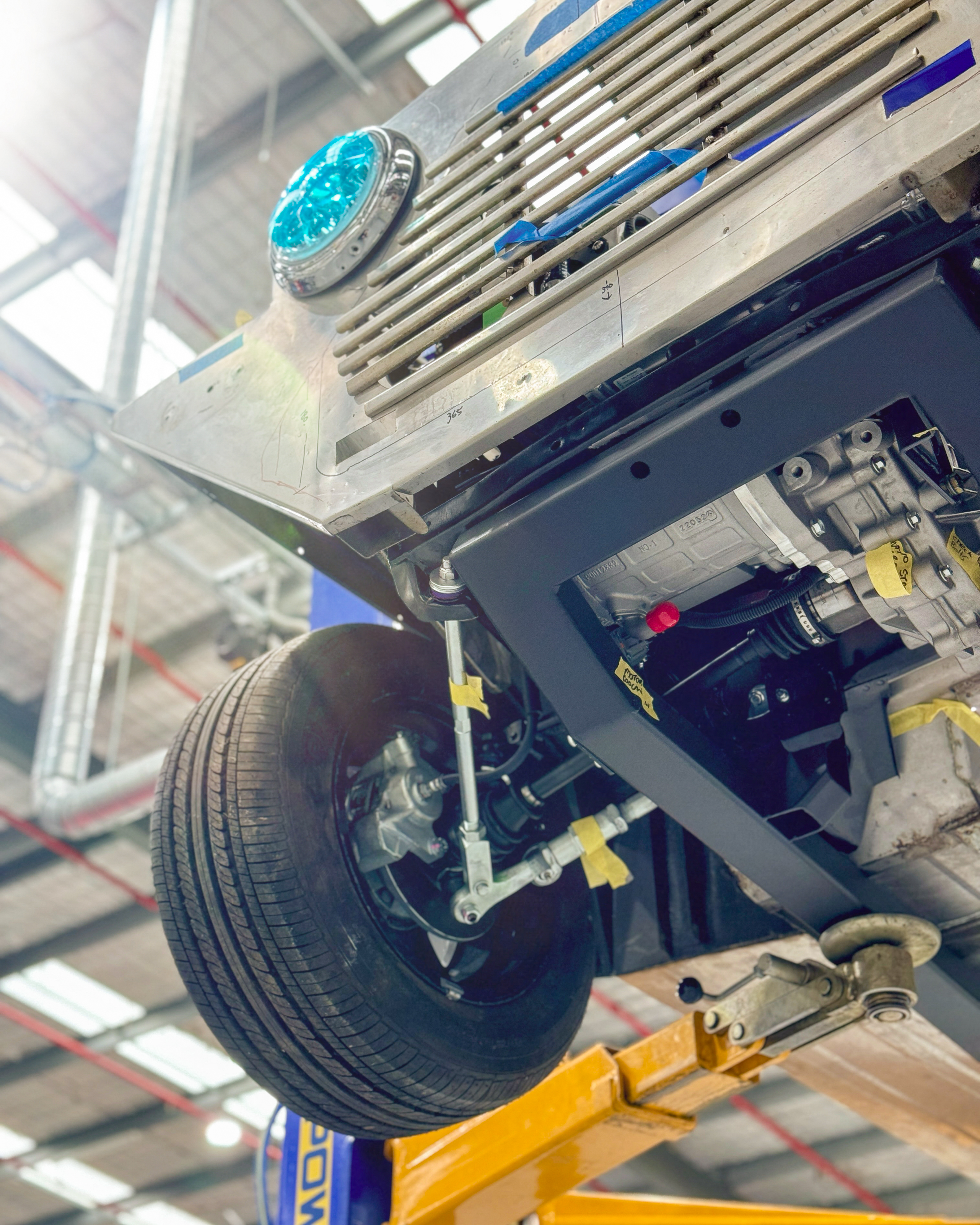 Low-angle view looking up at the front suspension and battery tray from beneath the raised Moke on a hoist, with the headlight visible above and the lift arm in frame.