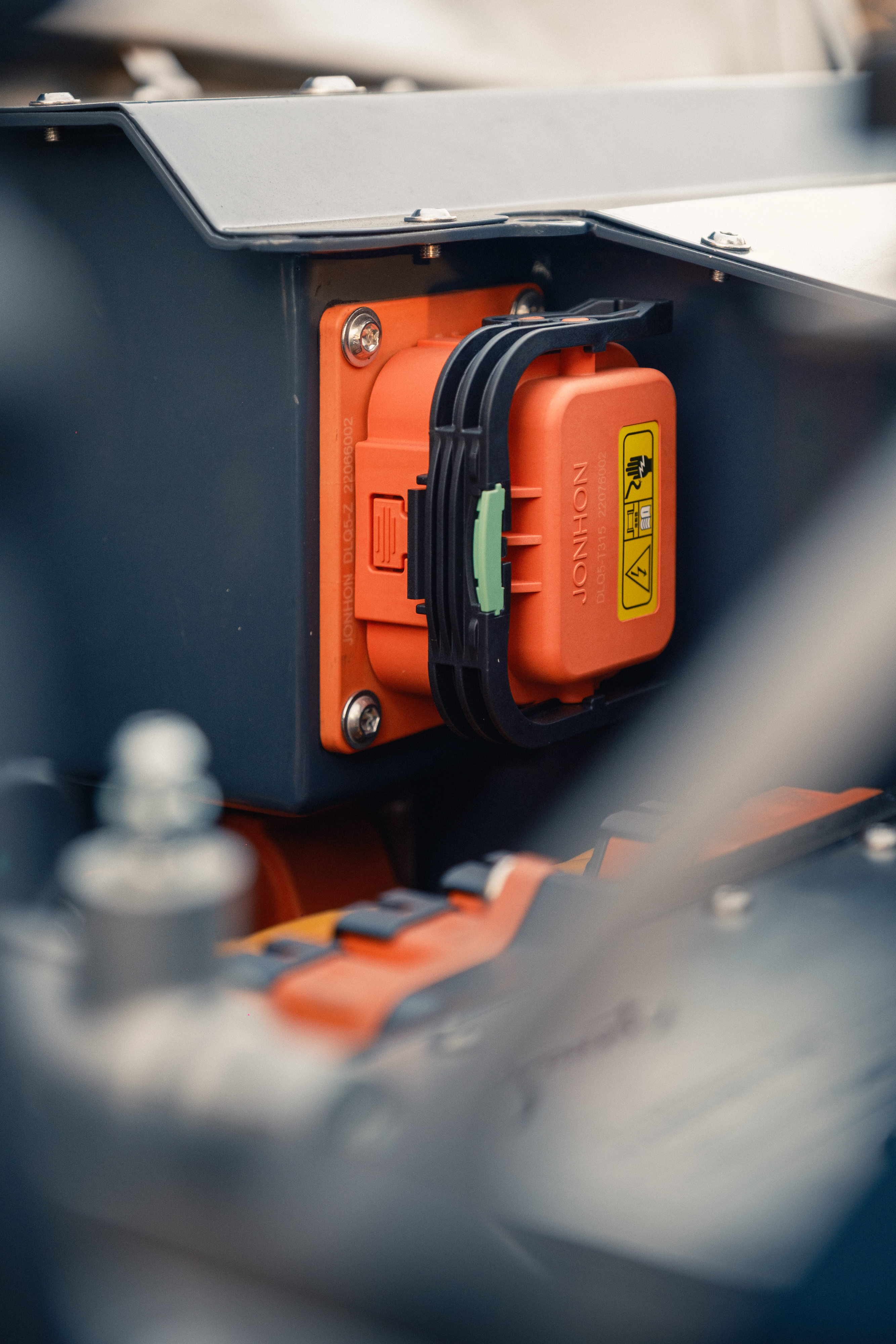 Close-up of the orange high-voltage Jonhon battery connector locked into a dark enclosure, with warning labels visible and additional orange HV cables below.