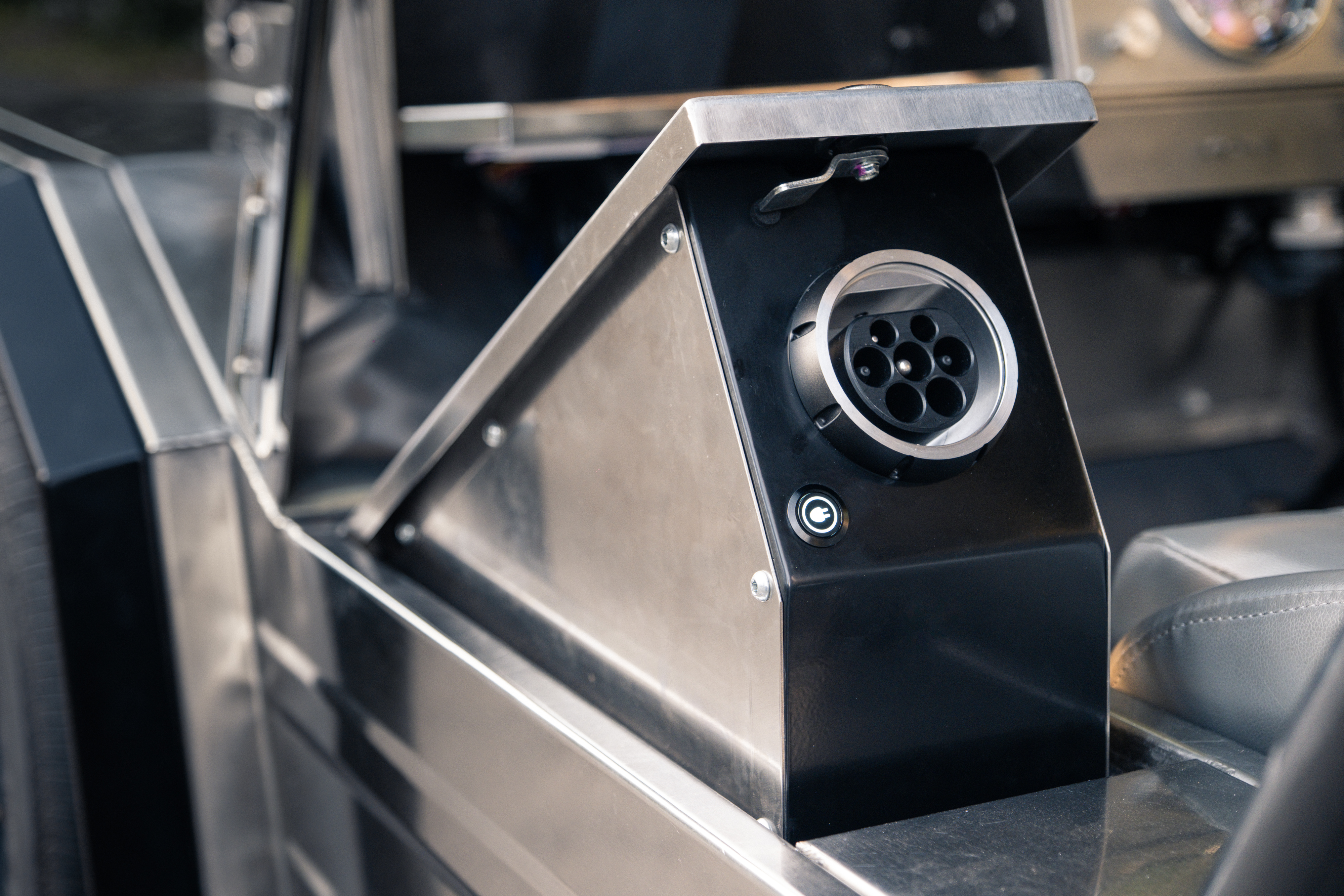 CCS2 fast-charge port revealed behind an open side-pod storage lid, showing the brushed stainless housing and a small illuminated charge-status indicator below.