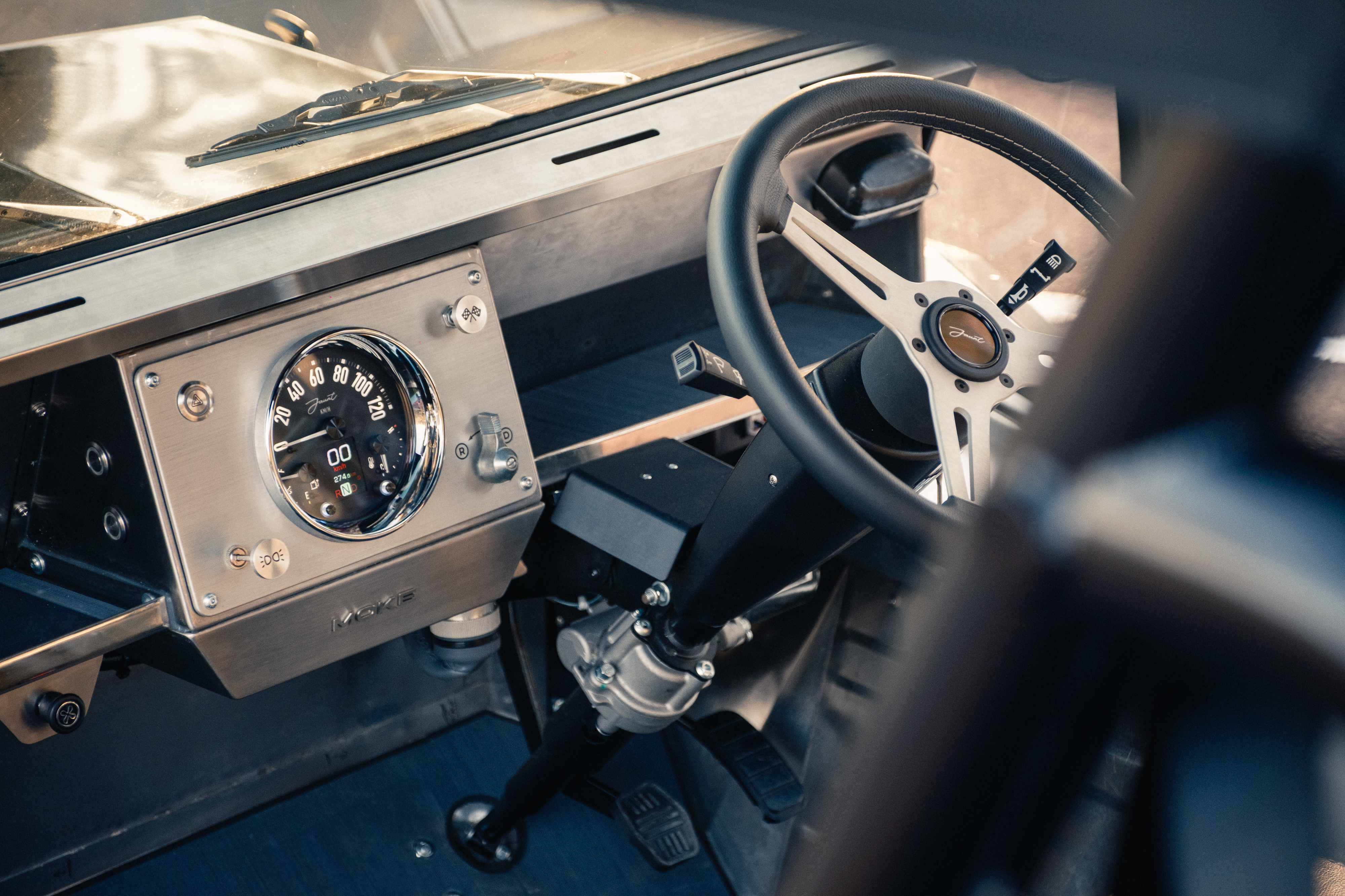 Driver's-eye view of the brushed stainless dashboard with the Jaunt analogue speedo, Moke-branded panel, and three-spoke steering wheel with Jaunt centre badge.