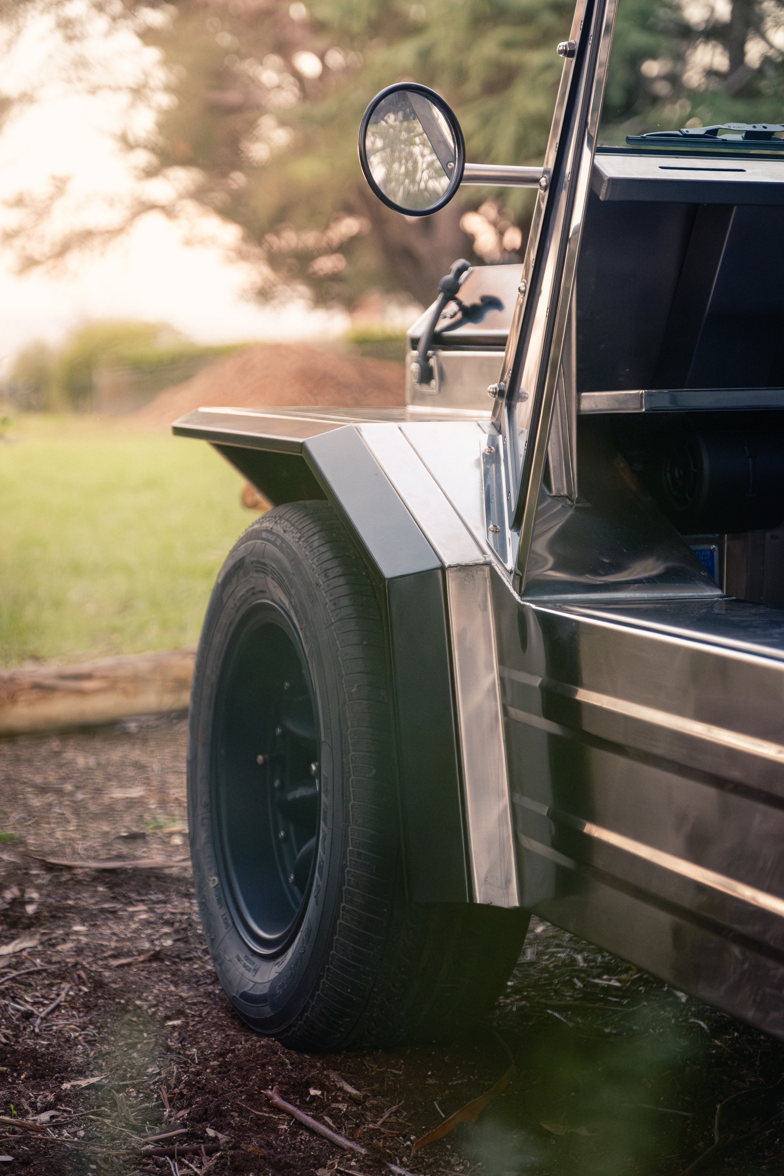 Low side-on view of the front guard and wheel in warm sunset light, polished stainless body catching the glow with eucalyptus trees soft in the background.