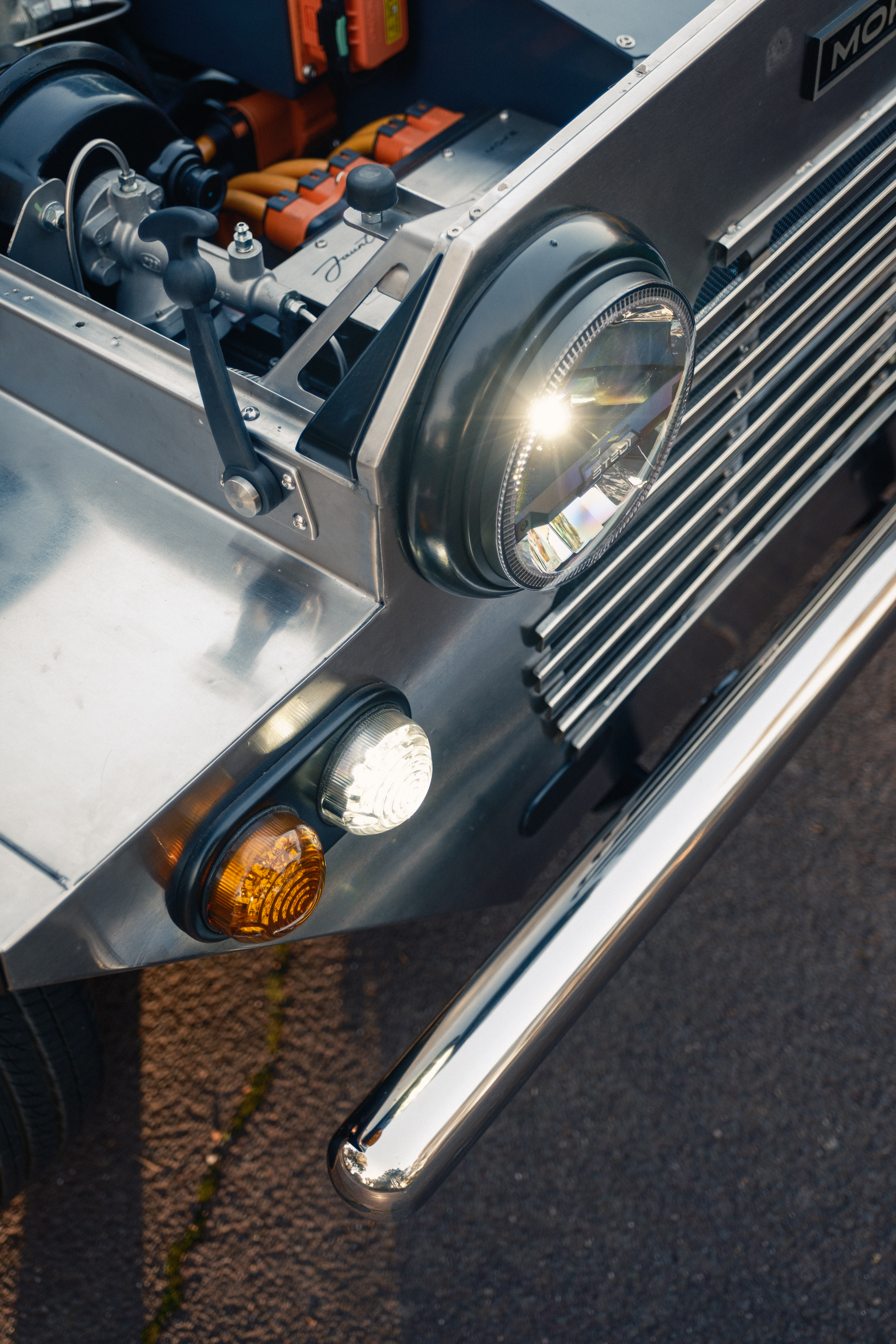 Front corner detail with the headlight on and EV bay open, revealing orange cables and the Jaunt-branded inverter bracket, with the stainless bumper tube across the bottom.
