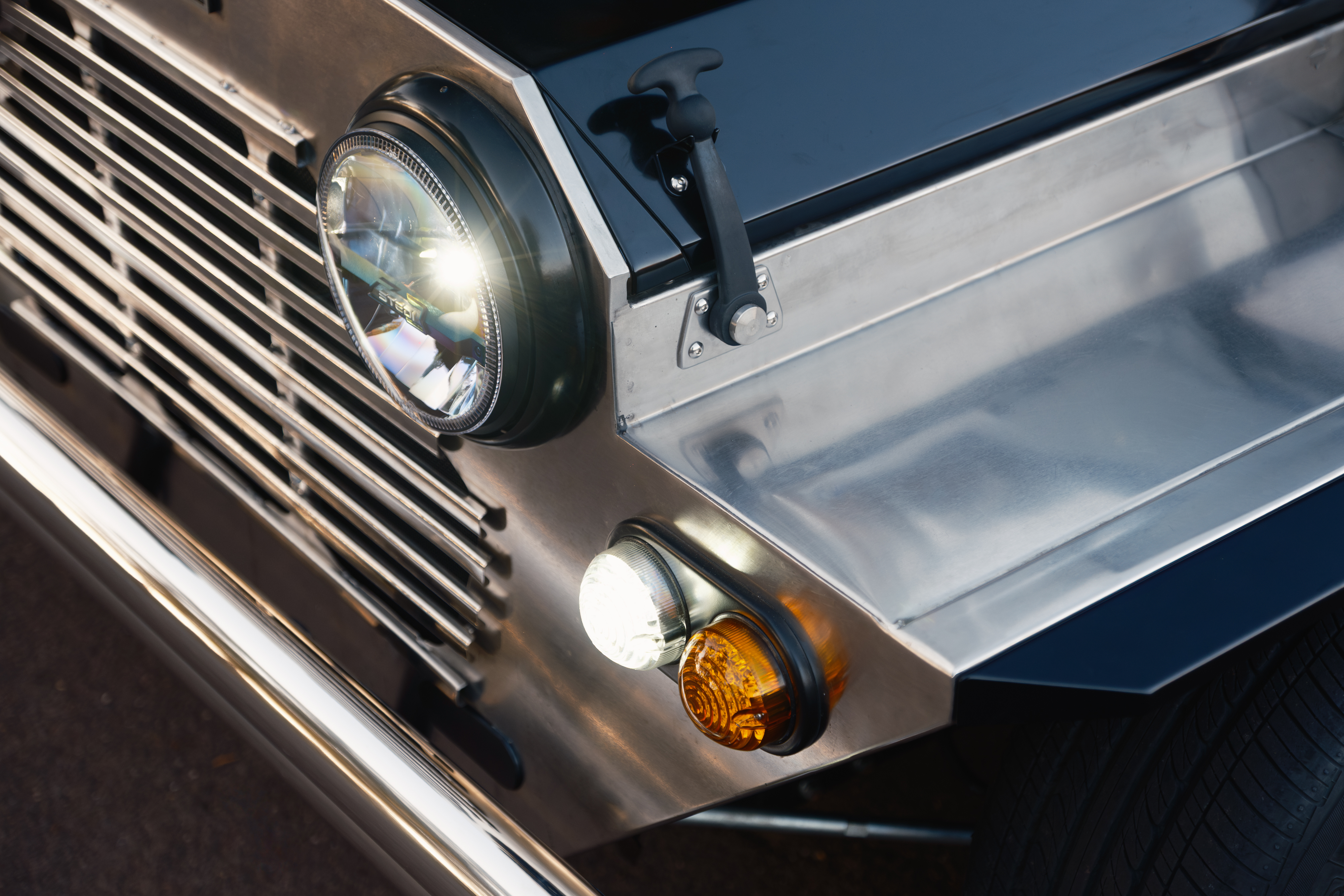 Close-up of the front corner showing the round LED headlight glowing, the amber indicator beside it, and the satin-black bonnet edge meeting the polished stainless guard.