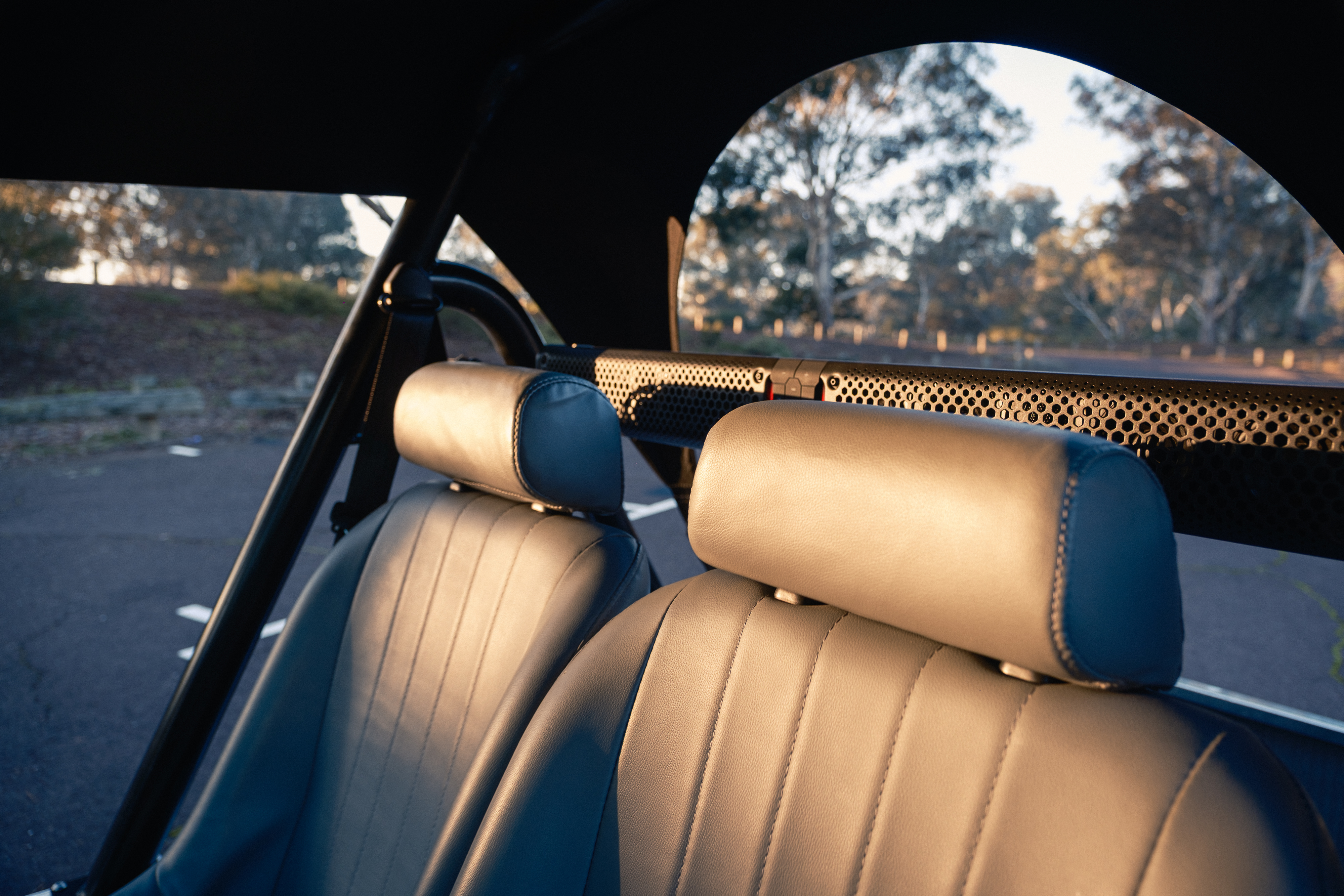 Interior view from behind looking forward, showing cream leather seats and the perforated speaker hoop, with a eucalyptus bush setting visible through the windscreen arch.
