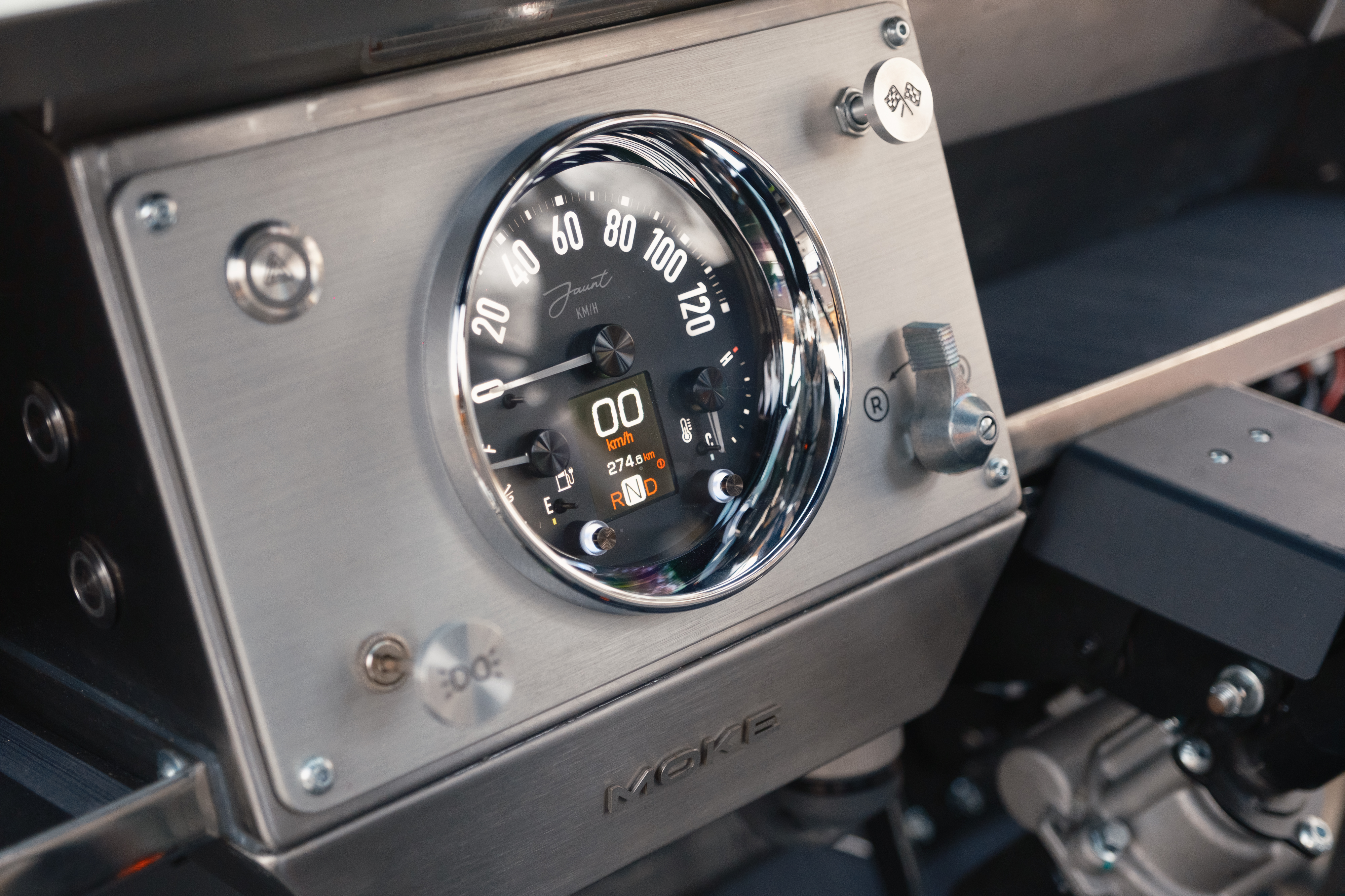 Macro view of the Jaunt-branded analogue speedometer on the brushed stainless dash panel, with the embedded digital display showing RND gear selector and odometer.