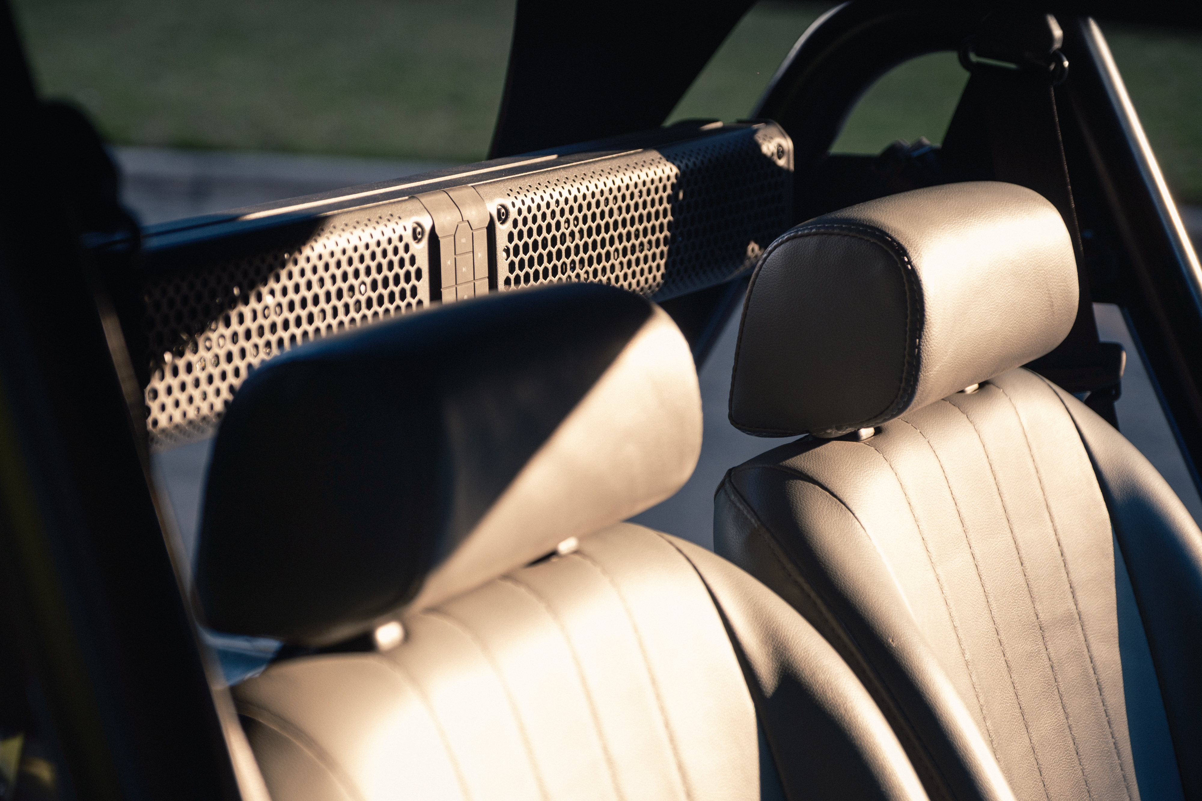 Close detail of the perforated stainless speaker grille integrated into the black tubular seat-belt hoop, with cream leather headrests in warm afternoon light.