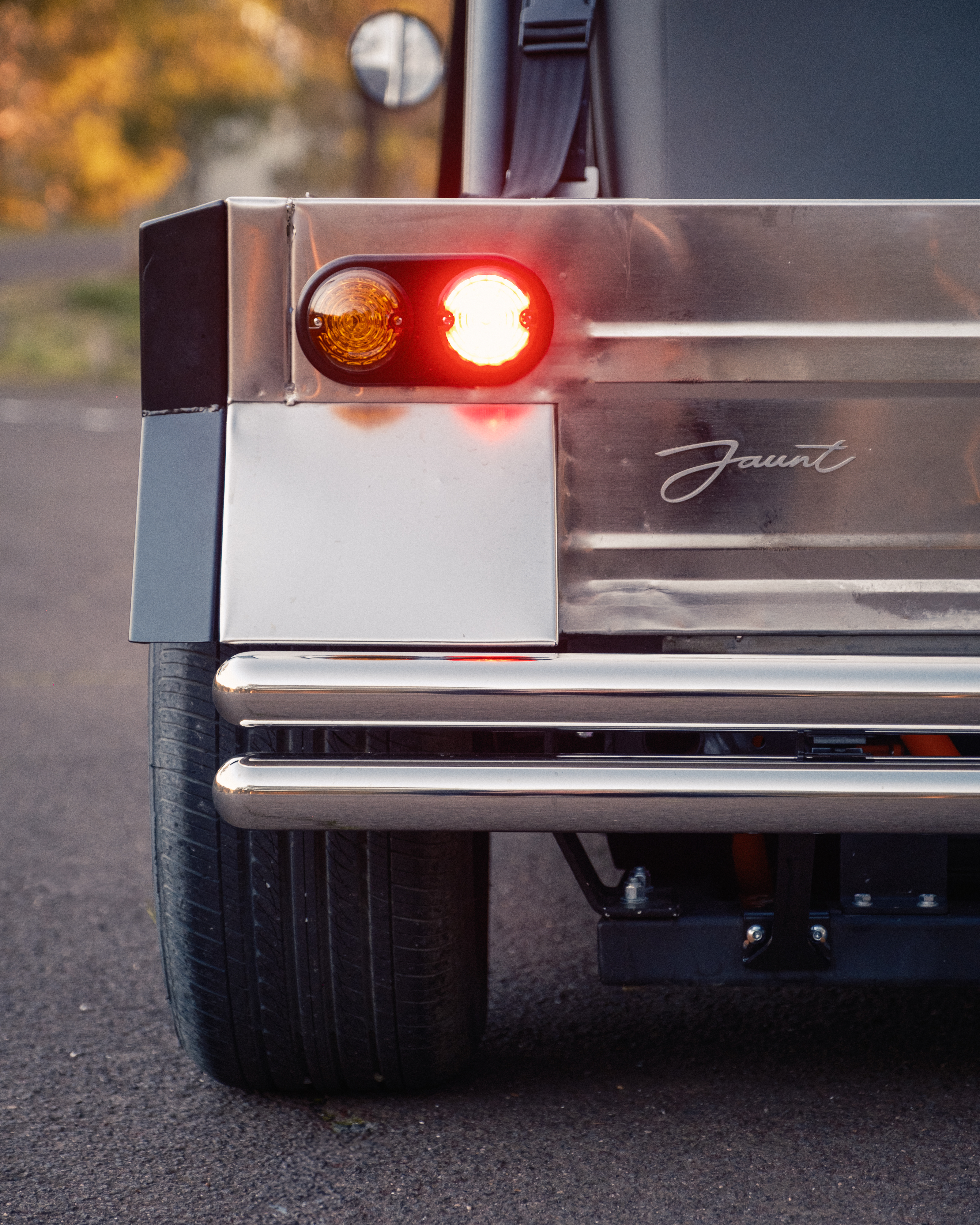 Tight rear detail showing the round LED taillight illuminated red beside the Jaunt script badge on the polished stainless body, with autumn foliage blurred behind.
