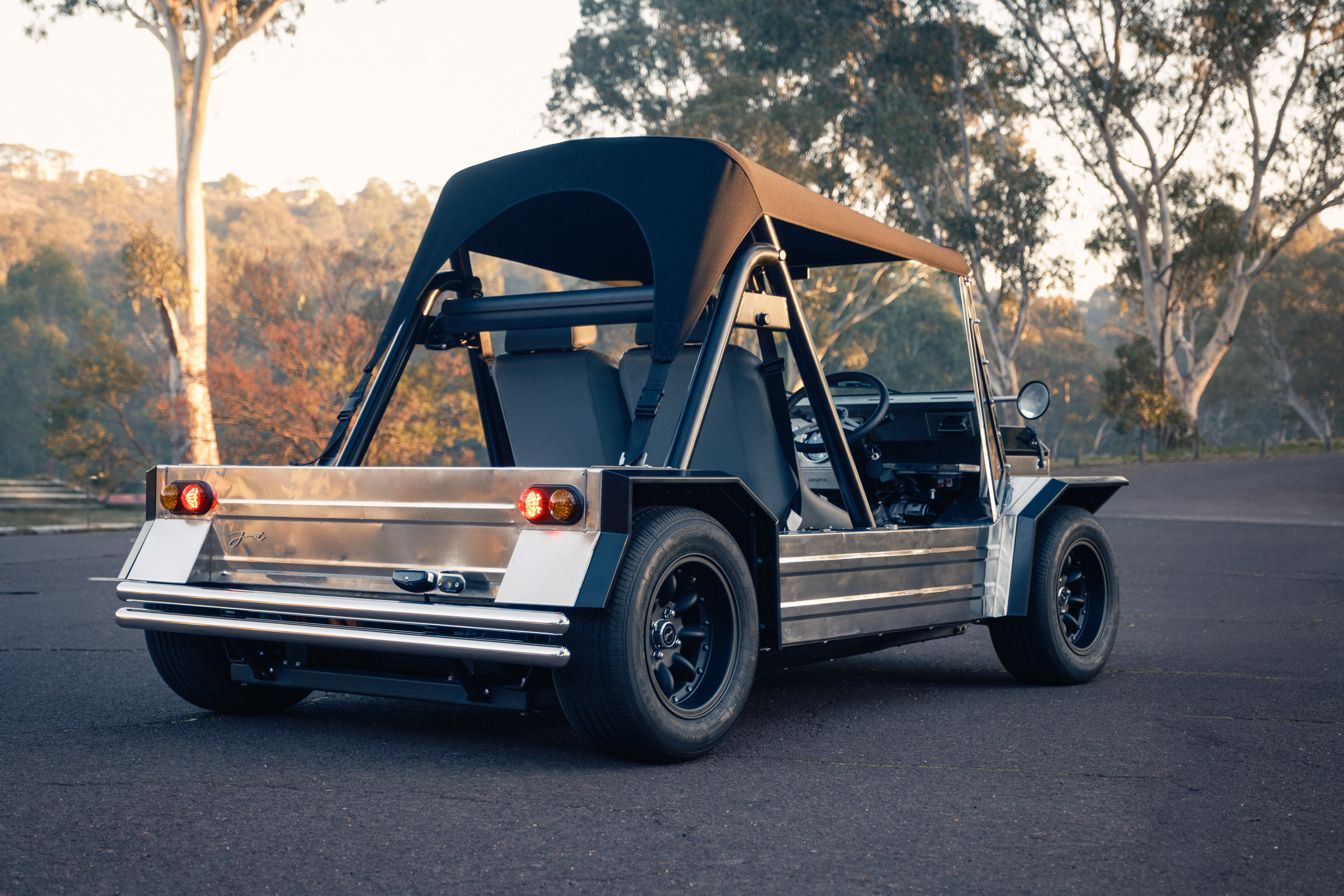 Three-quarter rear view of the Moke on a bush road at golden hour, taillights lit, with a canvas bikini top up and eucalyptus canopy glowing behind.