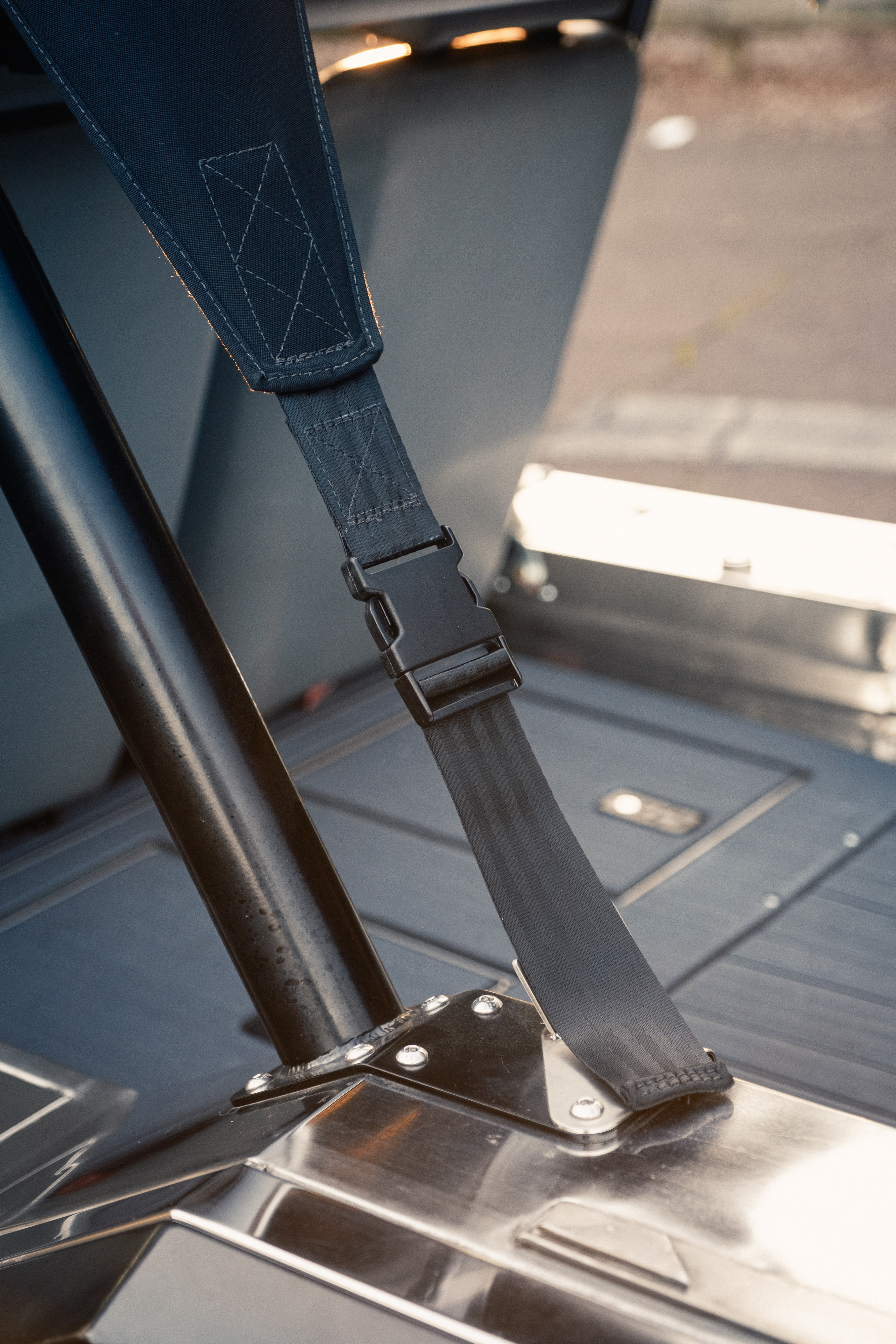Detail of the black tubular seat-belt hoop post and retractor strap anchored to the polished stainless floor, with the rear tub and sun-lit ground visible behind.