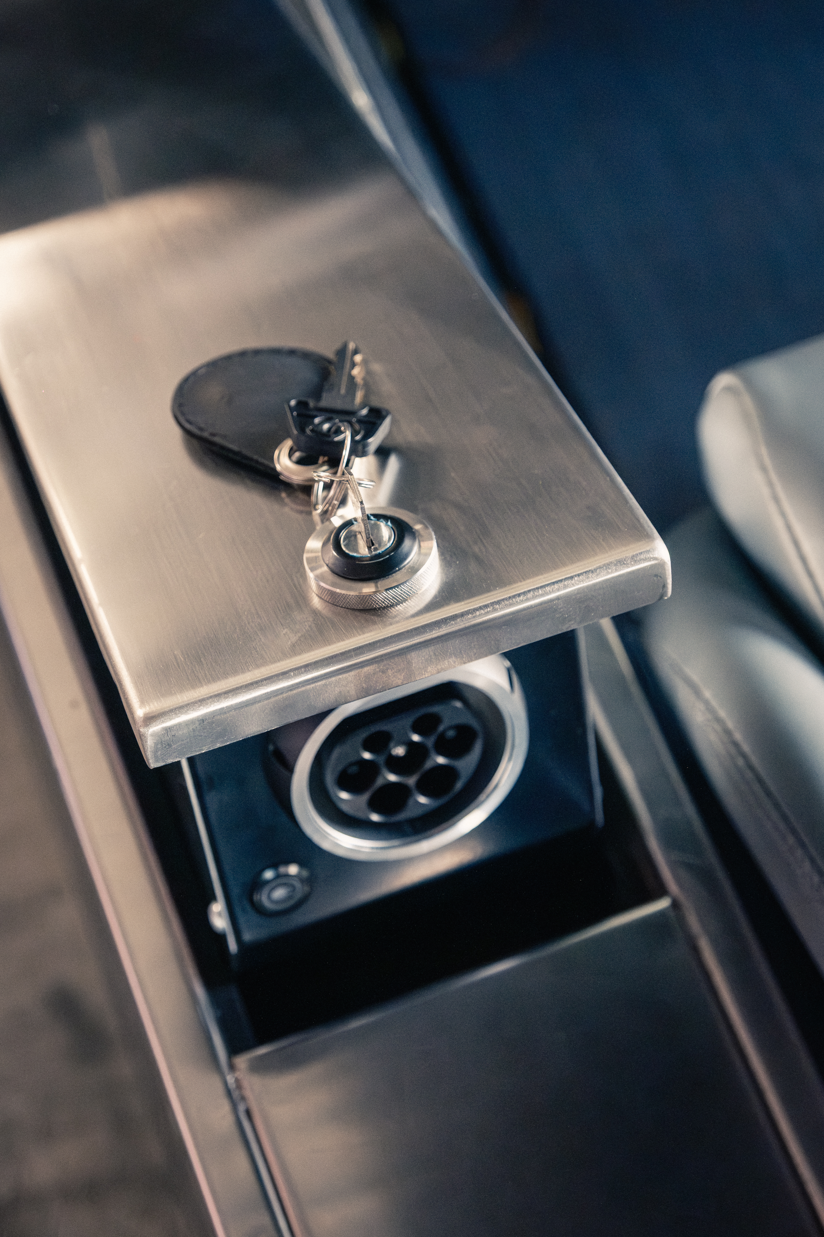 Top-down detail of the open side-pod storage lid revealing the CCS2 charge port below, with a leather key fob and keys resting on the brushed stainless lid surface.