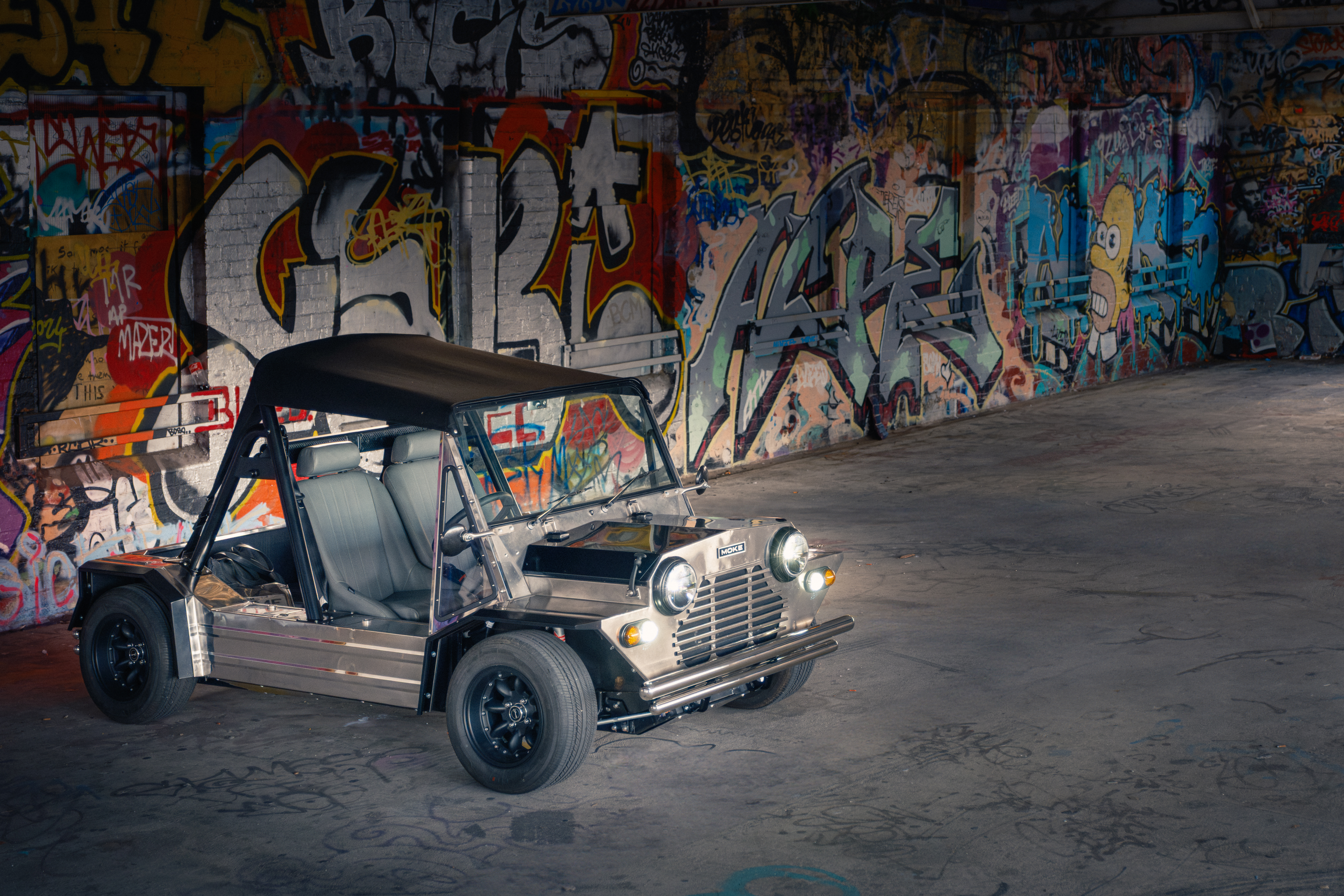 Front three-quarter angle inside the graffiti hall with headlights illuminated, the polished stainless body picking up the surrounding colourful murals as reflections.