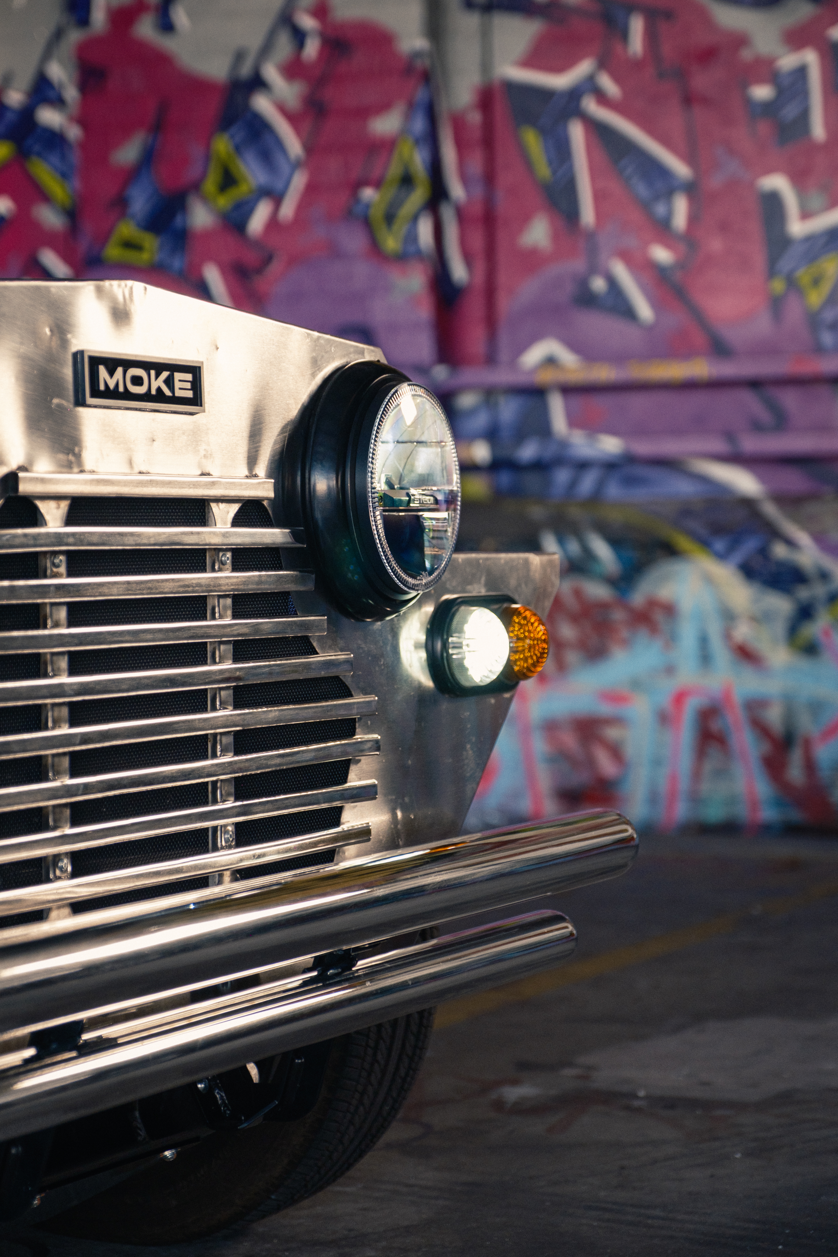 Close detail of the polished stainless front grille with the MOKE badge and round headlight, a pink-and-blue graffiti mural filling the background.