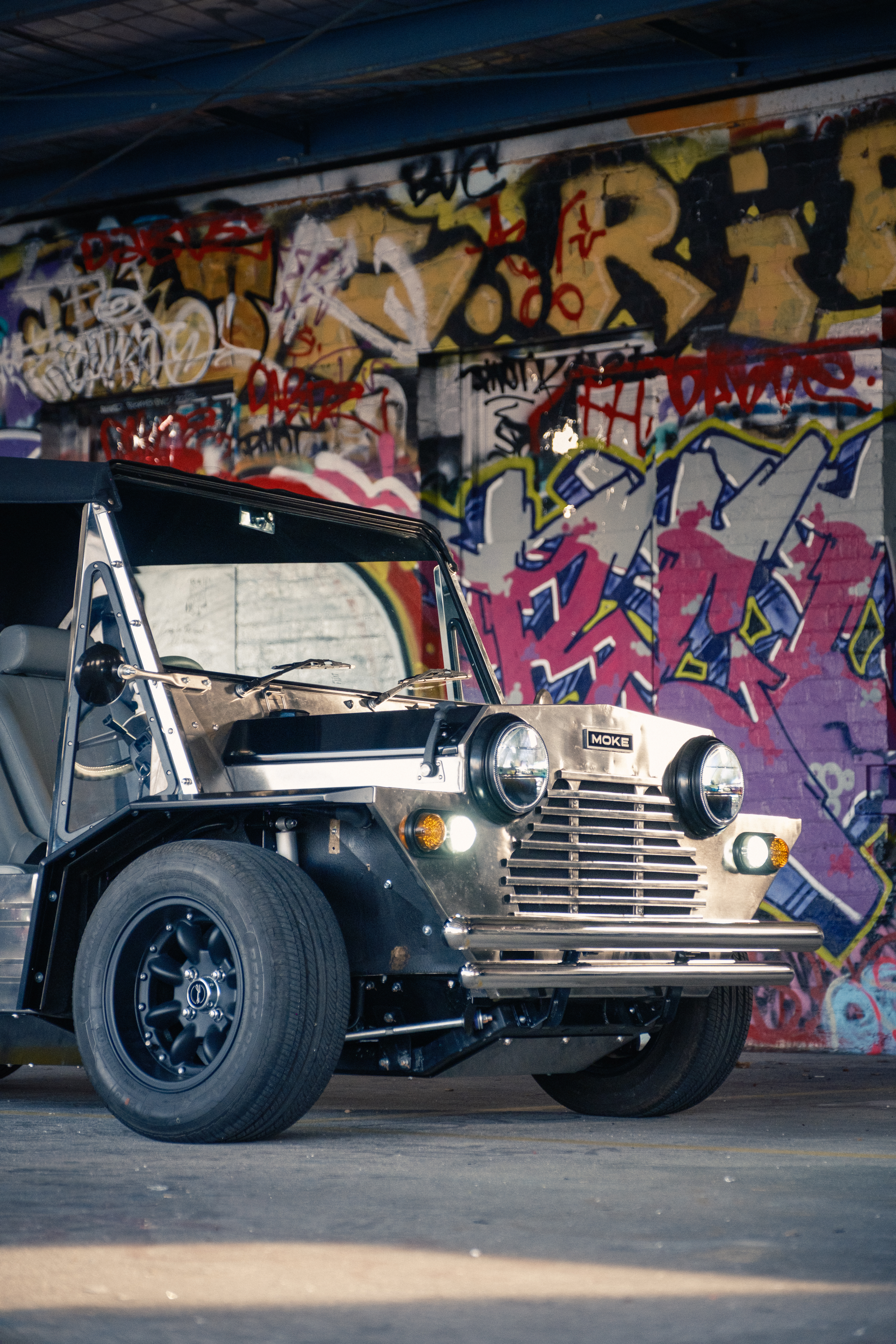 Three-quarter front view of the stainless Moke parked against a brightly painted graffiti wall in an urban setting, with colourful lettering filling the background.