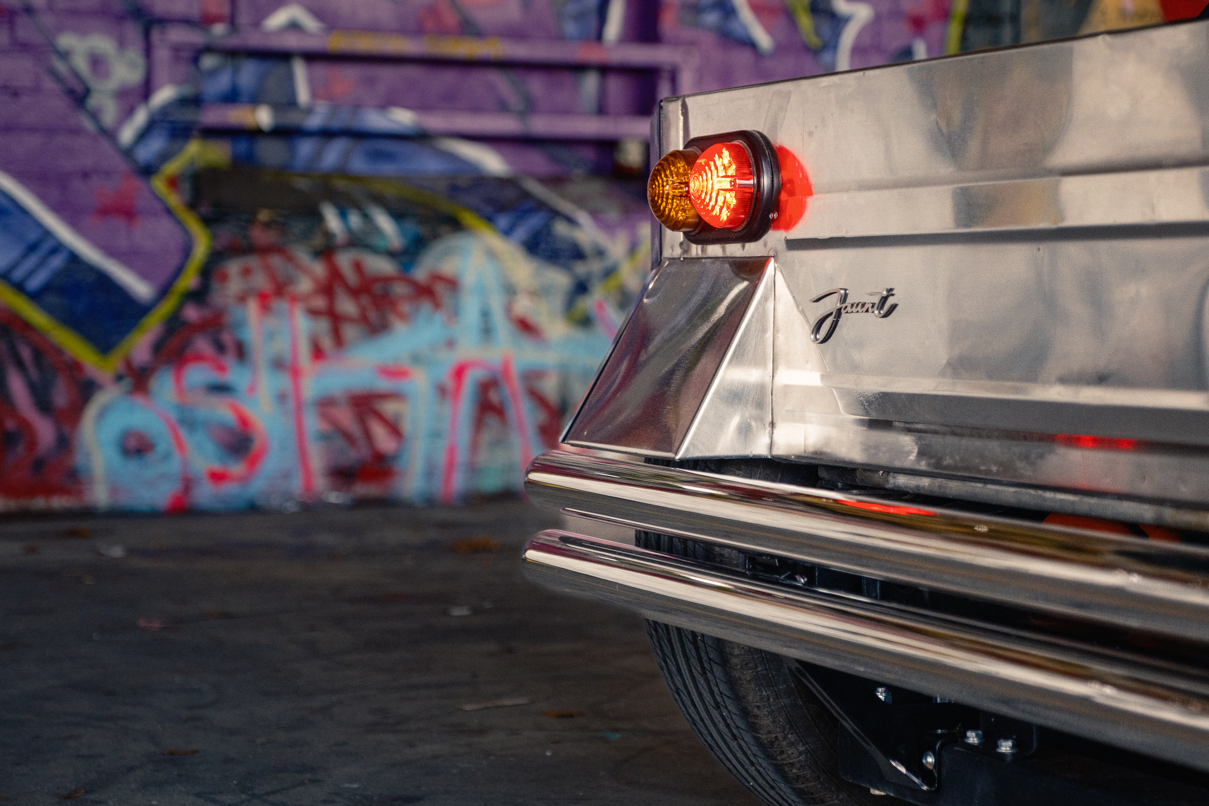 Rear detail showing the illuminated red taillight beside the Jaunt script badge on the polished body, with a blue-and-purple graffiti wall behind.