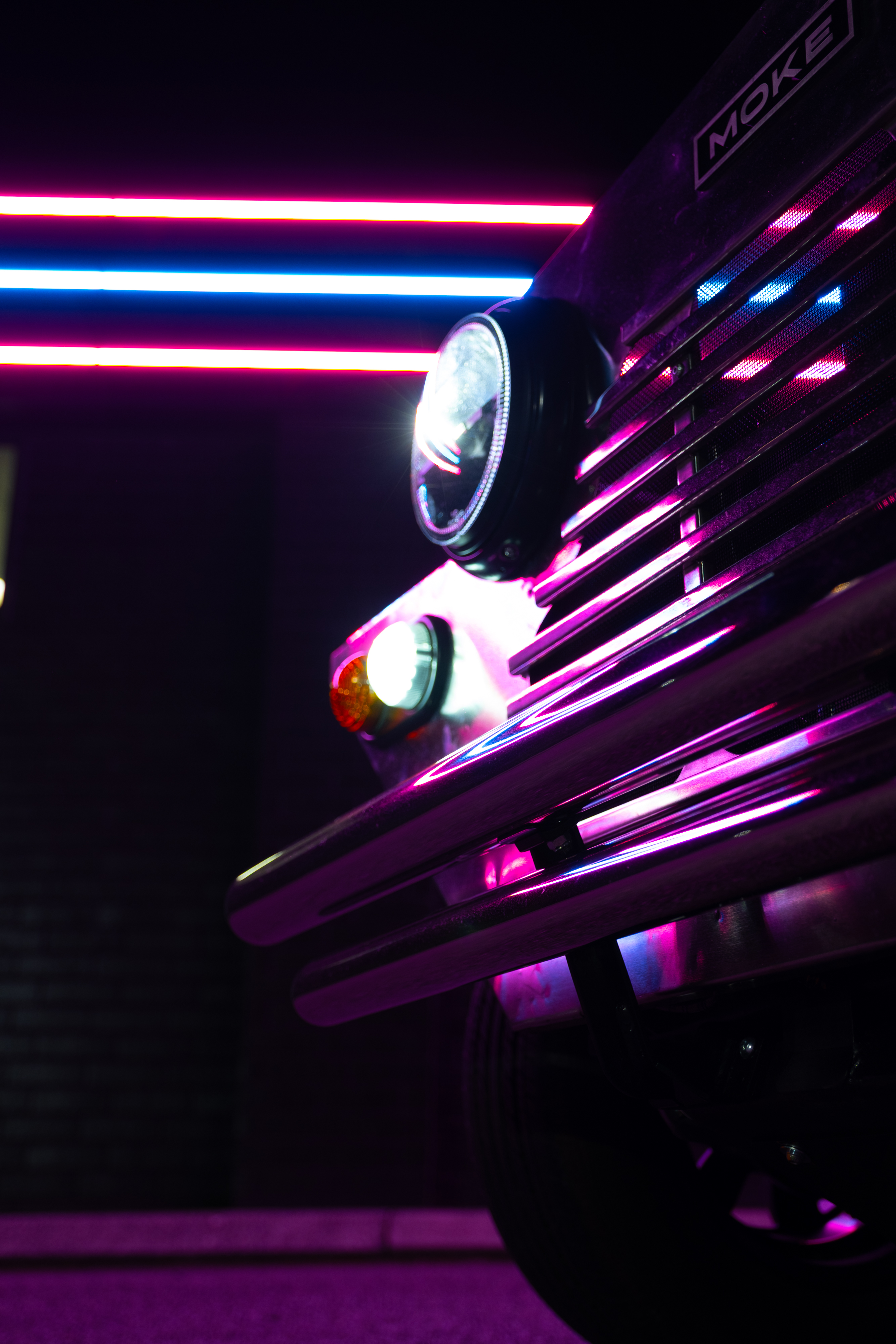 Dark low-angle shot of the front grille and headlight at night with parallel pink and blue neon strips running diagonally across the brick wall behind.