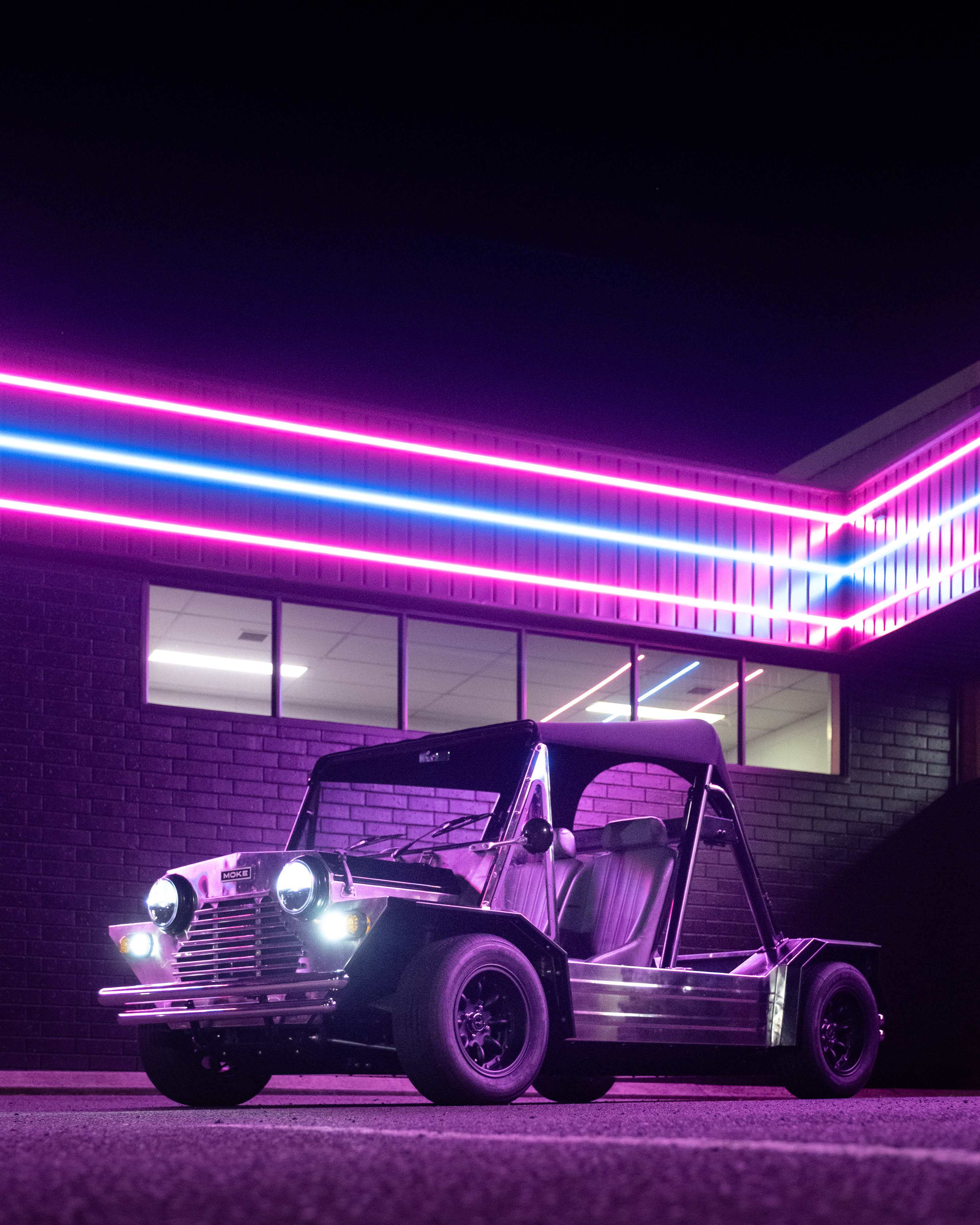Low front three-quarter view at night with headlights blazing and pink-blue LED strips above on a brick building, the body glowing deep violet from the ambient neon.