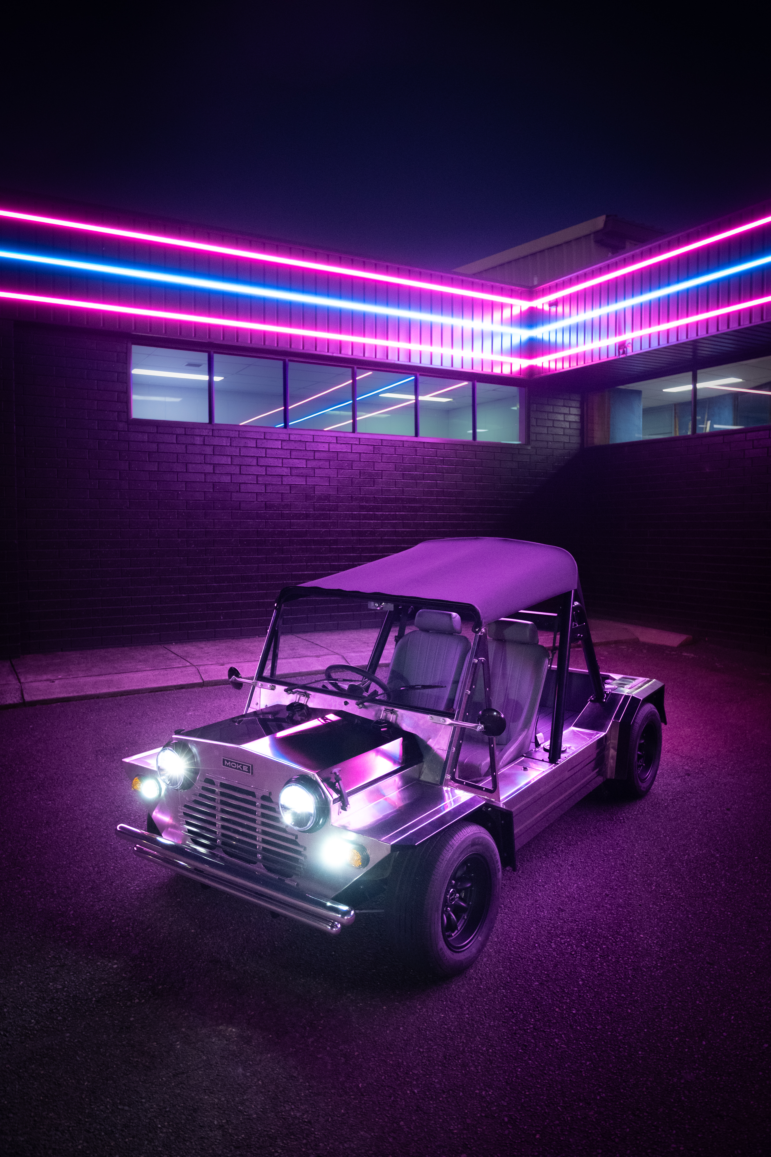Three-quarter front view of the Moke at night under a building wrapped with parallel pink and blue LED strips, the polished body glowing magenta.