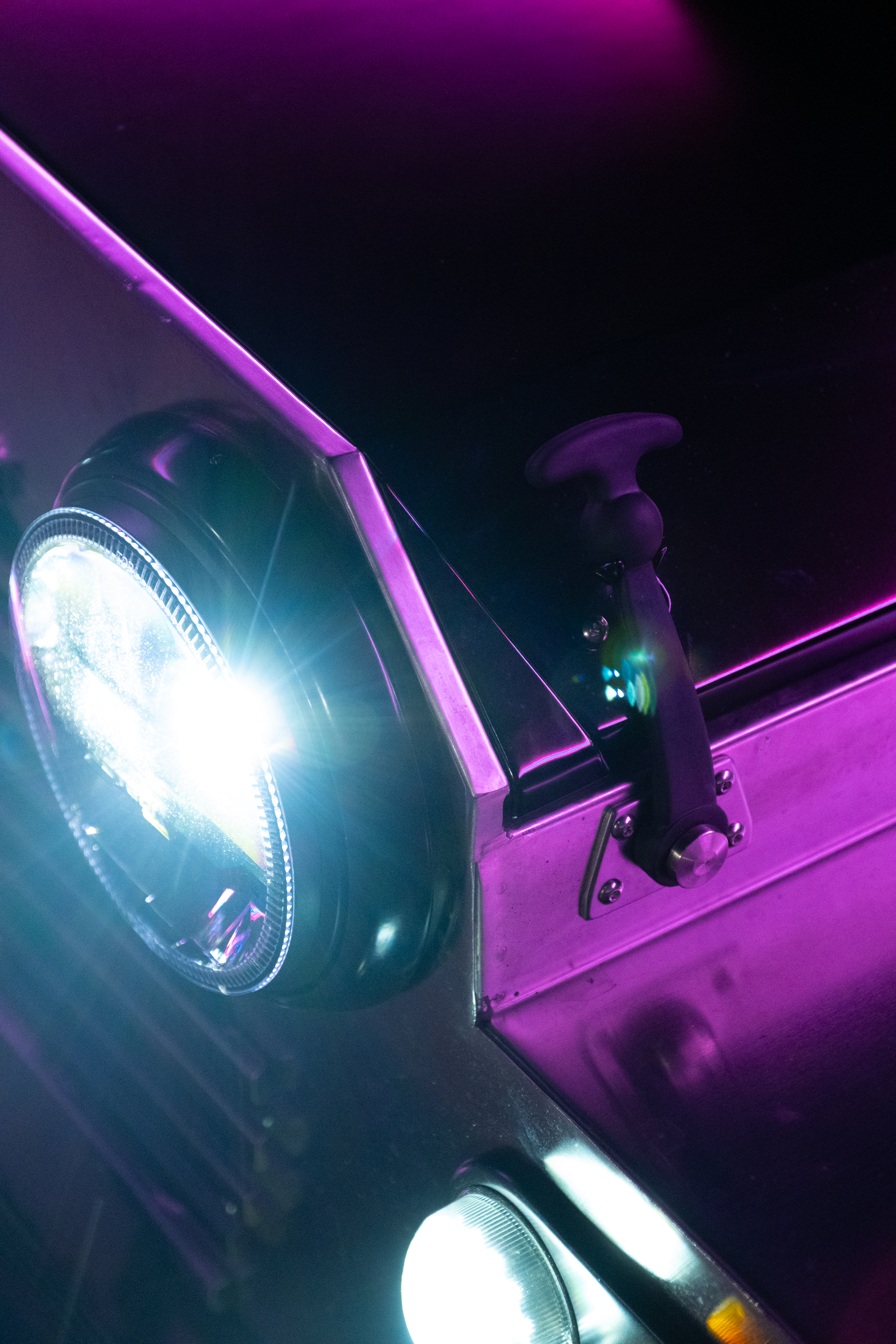 Low upward angle of the headlight blazing white against a magenta-bathed stainless panel at night, with a cyan-pink neon tube visible in the upper background.