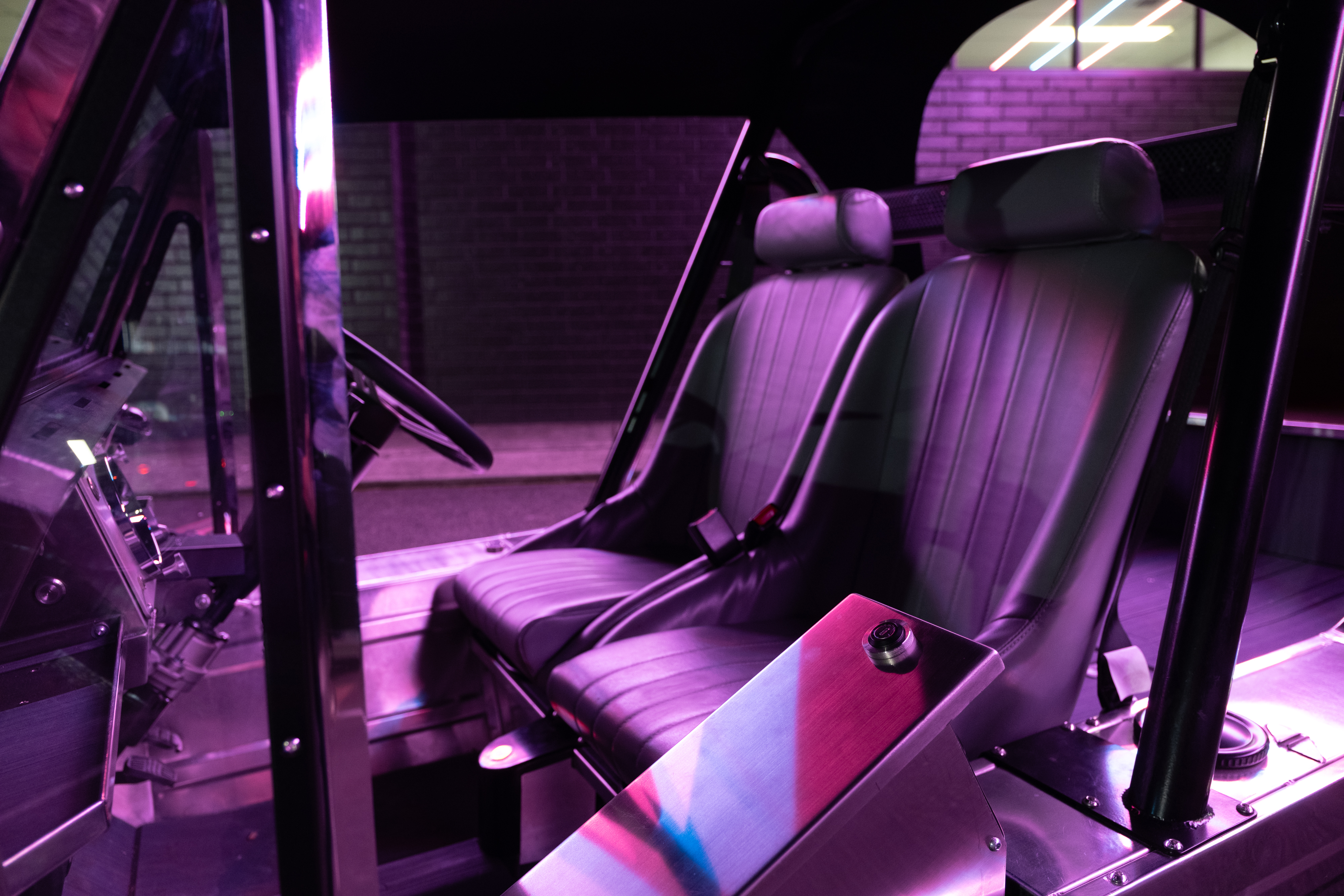 Interior view looking across the seats and console under pink neon ambient light, showing the leather upholstery and the brushed stainless tunnel console with power button.