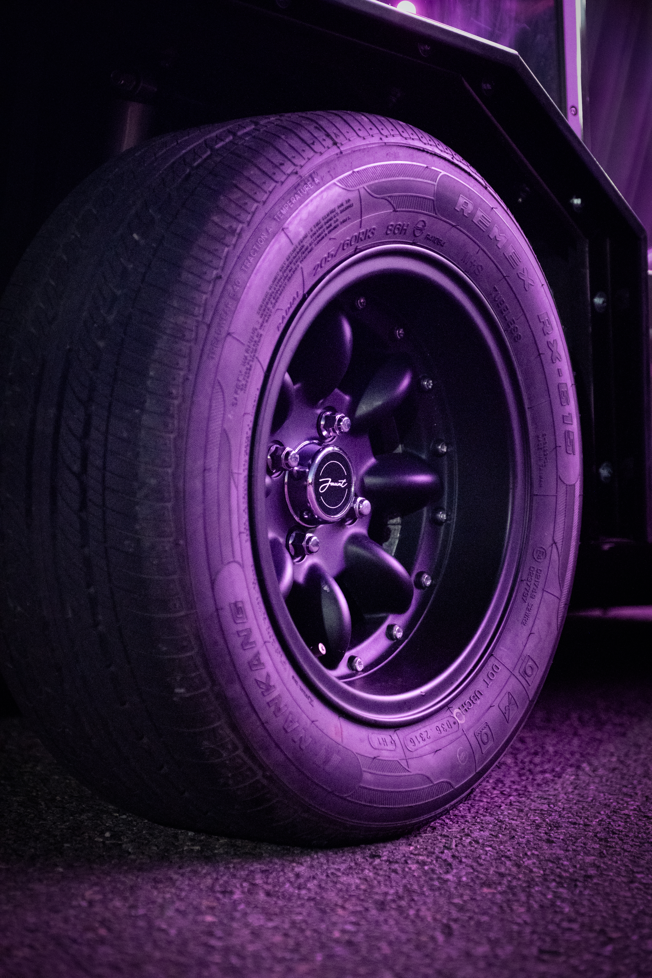 Close-up of the black alloy wheel with the Jaunt script centre cap, bathed in deep magenta neon light against a dark background.