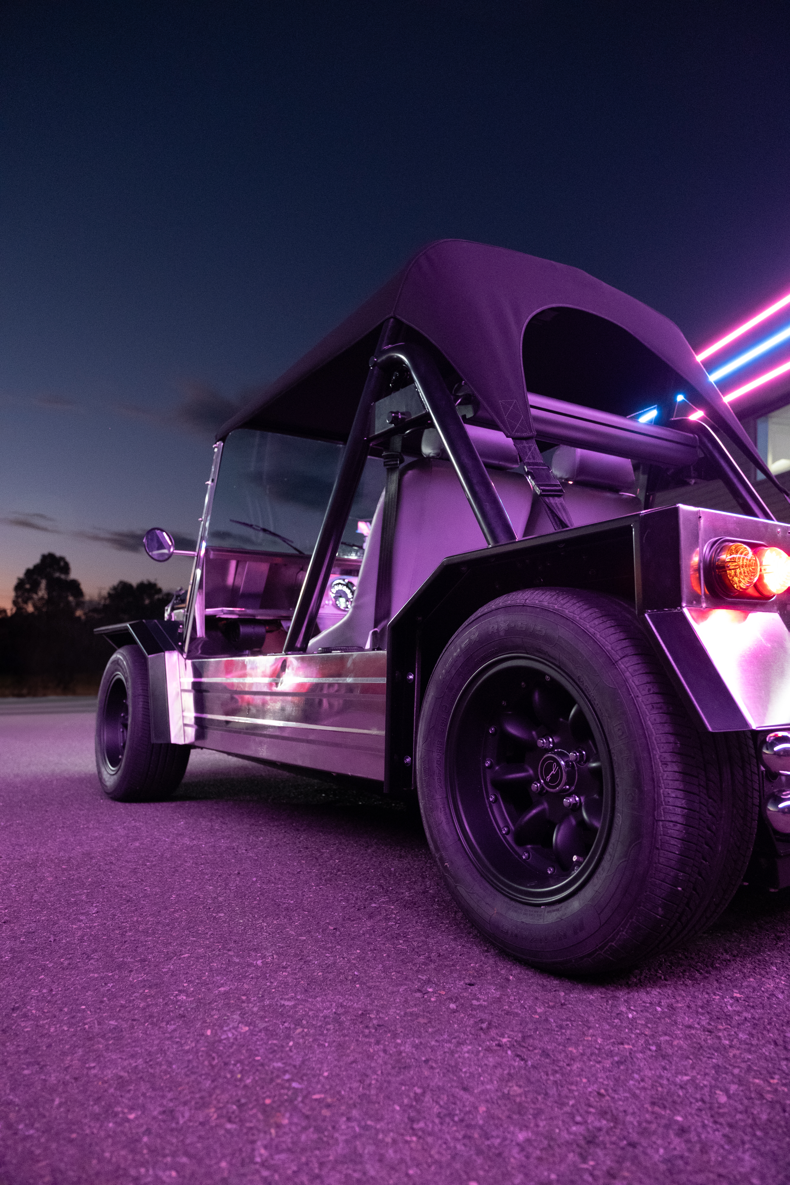 Rear three-quarter view at dusk with the deep blue twilight sky above and a pink neon strip running along the building roofline behind the Moke.