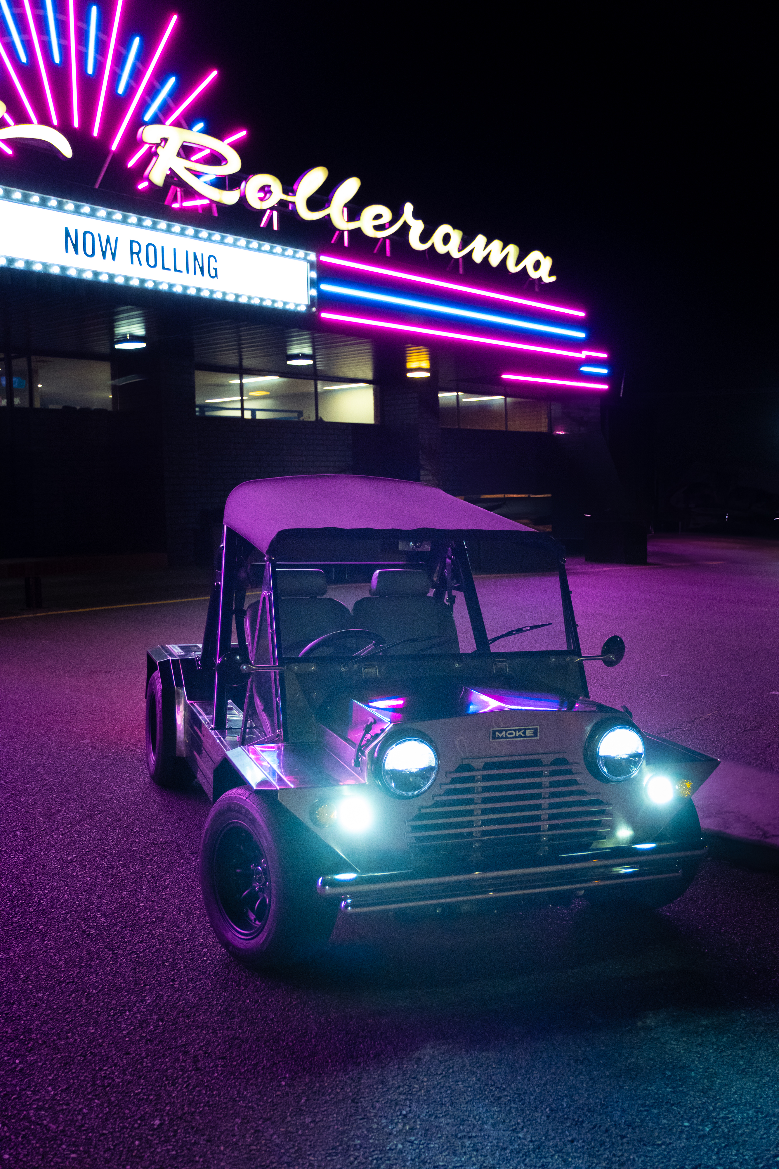 Front view of the Moke at night parked in front of the Rollerama roller-skating venue sign with its pink neon starburst and blue-and-pink LED roof strips.