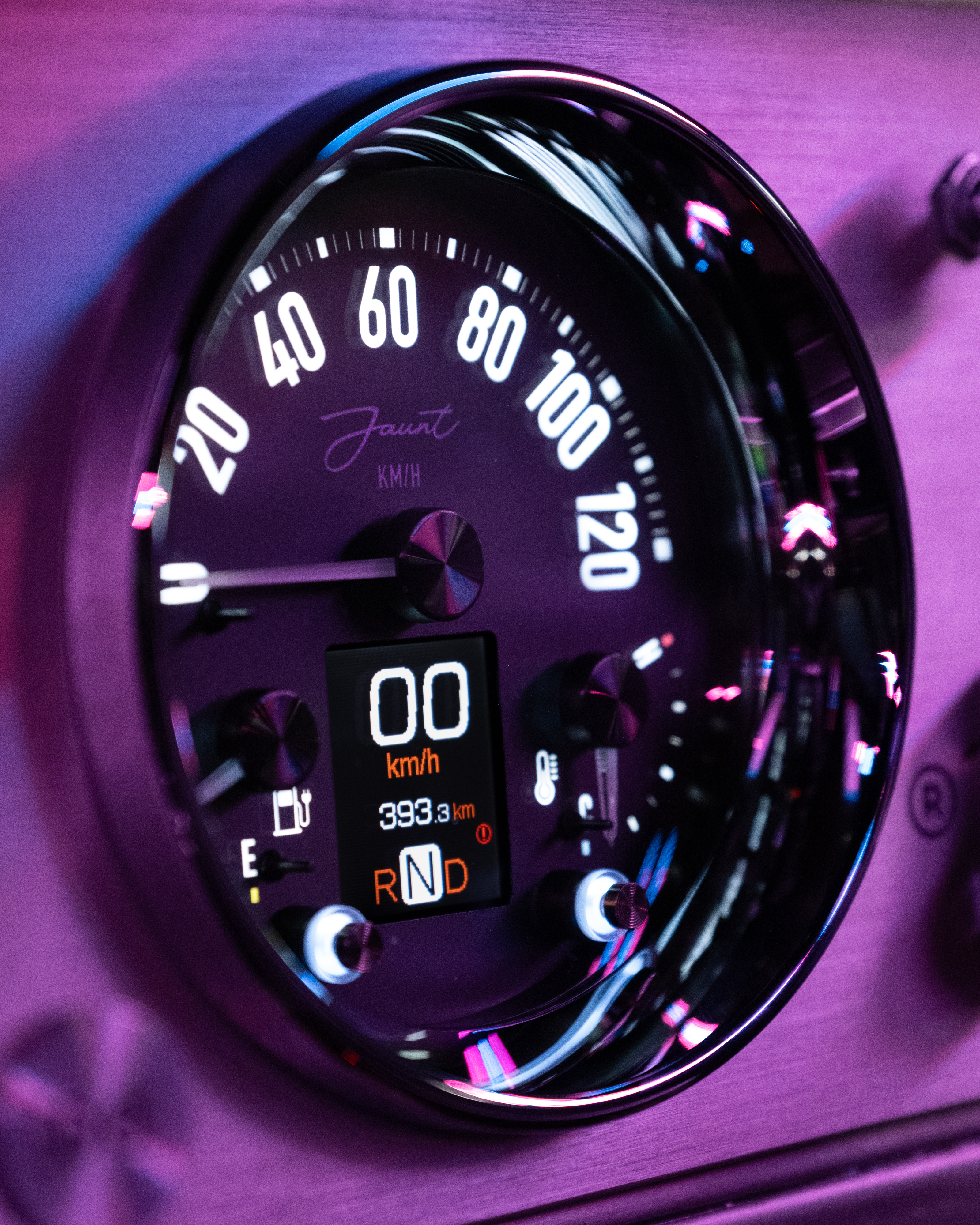 Extreme macro of the Jaunt analogue speedo face under pink neon, the chrome bezel reflecting surrounding lights and the digital display clearly showing RND mode.
