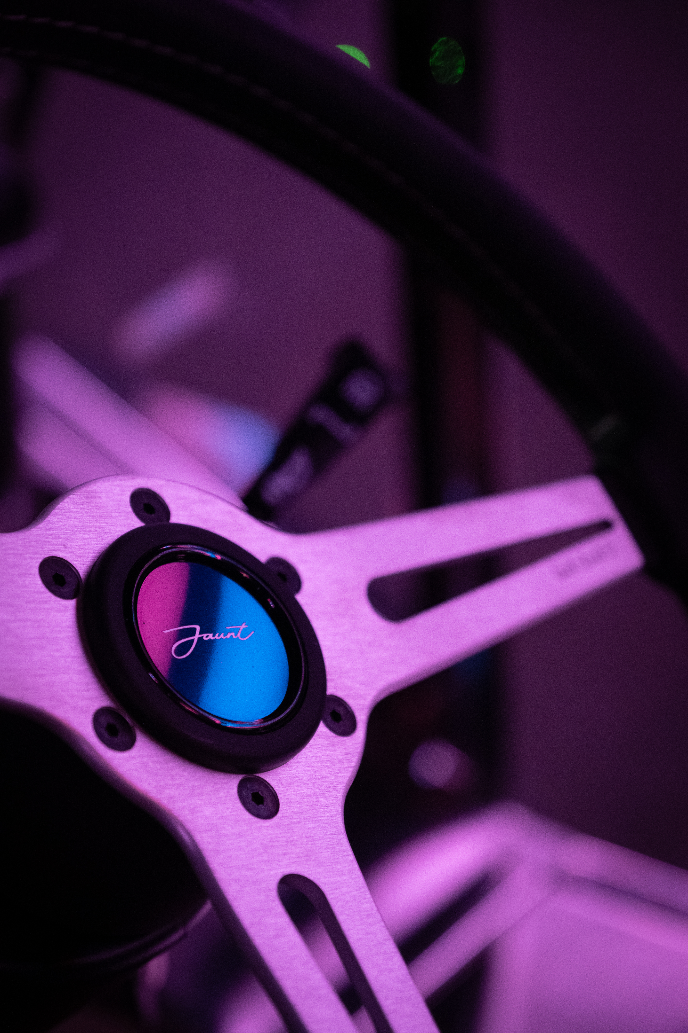 Close-up of the three-spoke brushed steering wheel and Jaunt centre badge under magenta neon, the badge face catching pink and blue reflections.