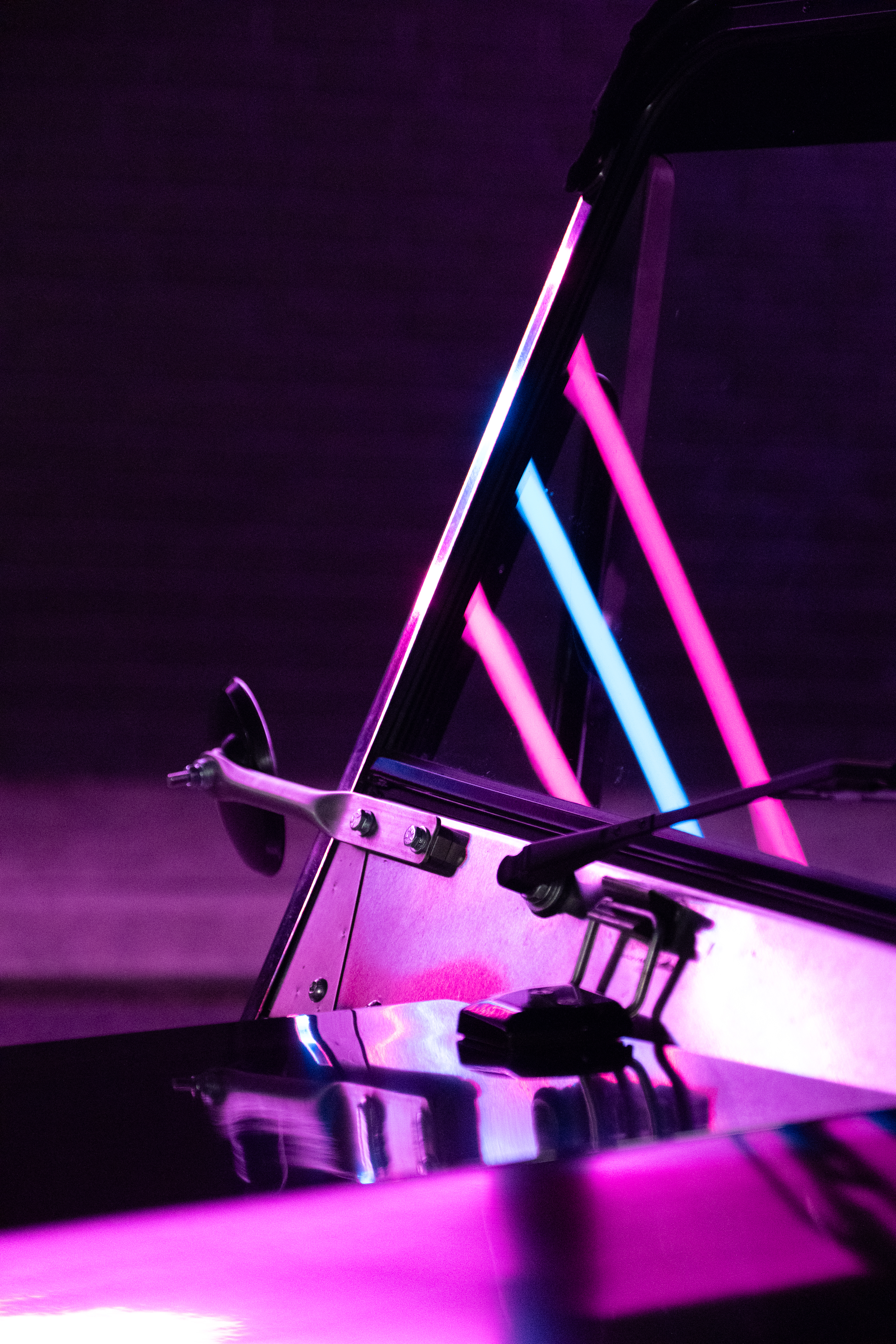 Side detail of the windscreen frame and seat-belt hoop reflecting pink and cyan neon strips at night, the polished stainless body acting as a mirror below.