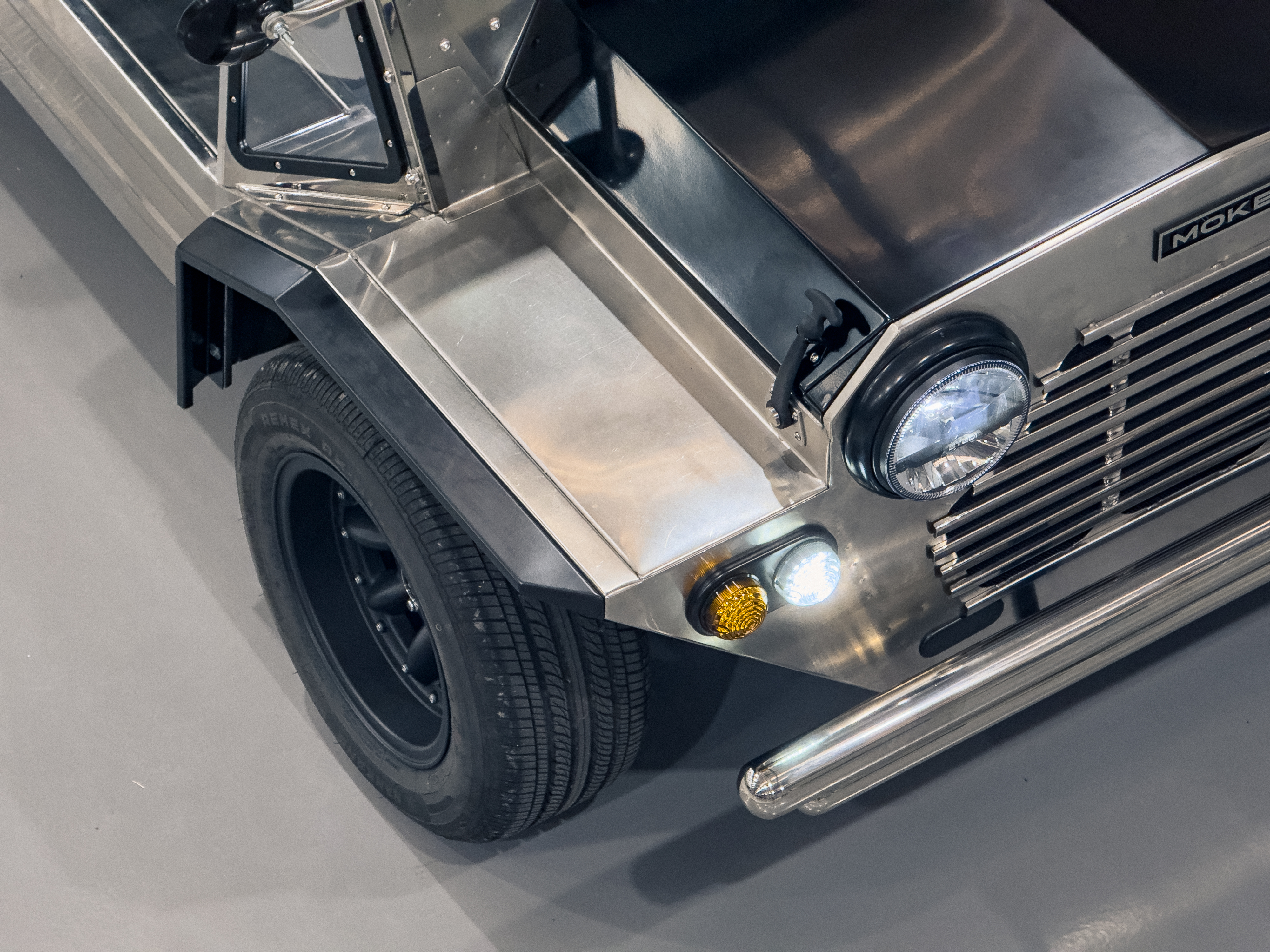 Overhead front-quarter view looking down at the headlight, guard, and indicator all illuminated, the polished stainless panels reflecting the workshop floor.