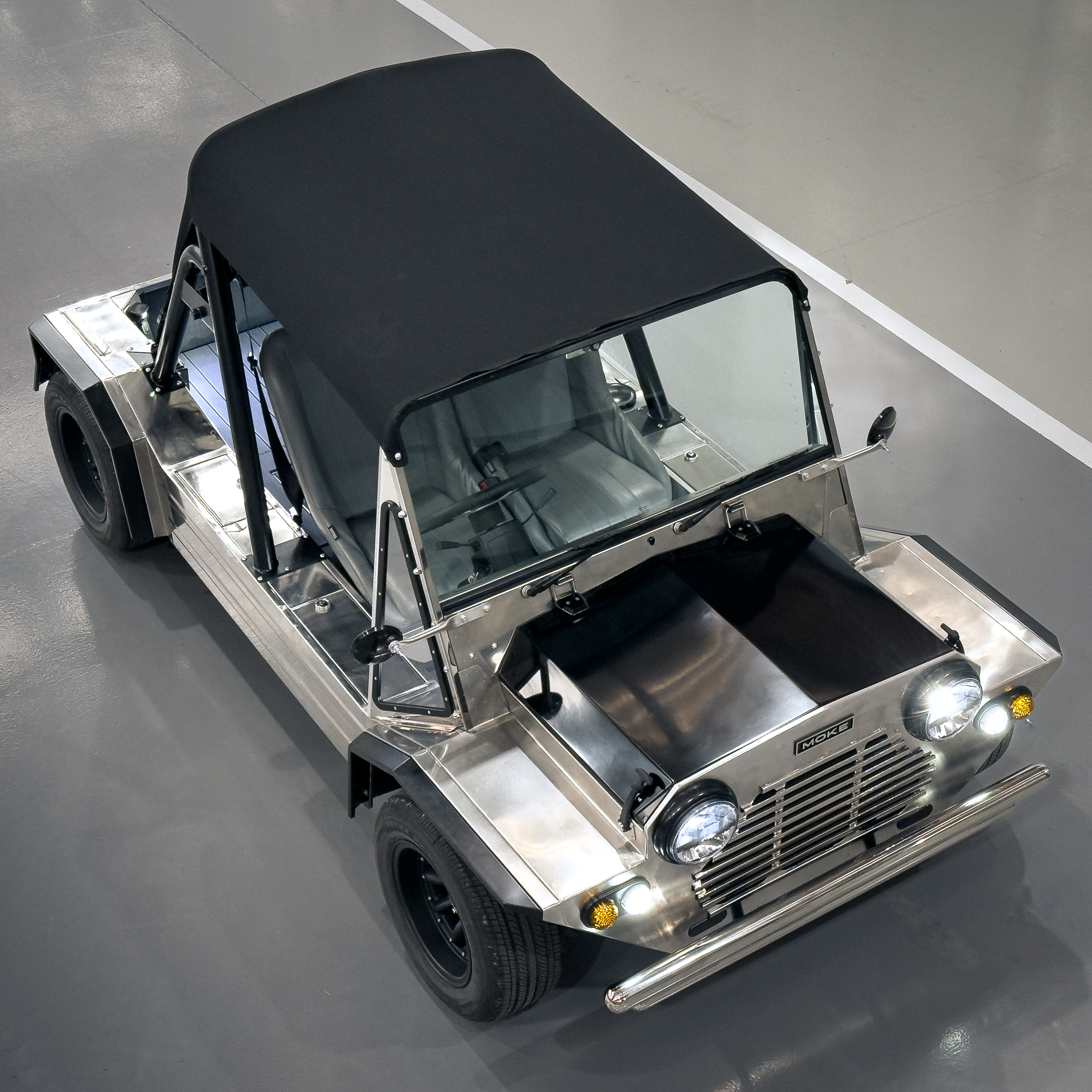 High overhead three-quarter front view of the Moke on the workshop floor with headlights and indicators on, showing the full black canvas roof and polished body.