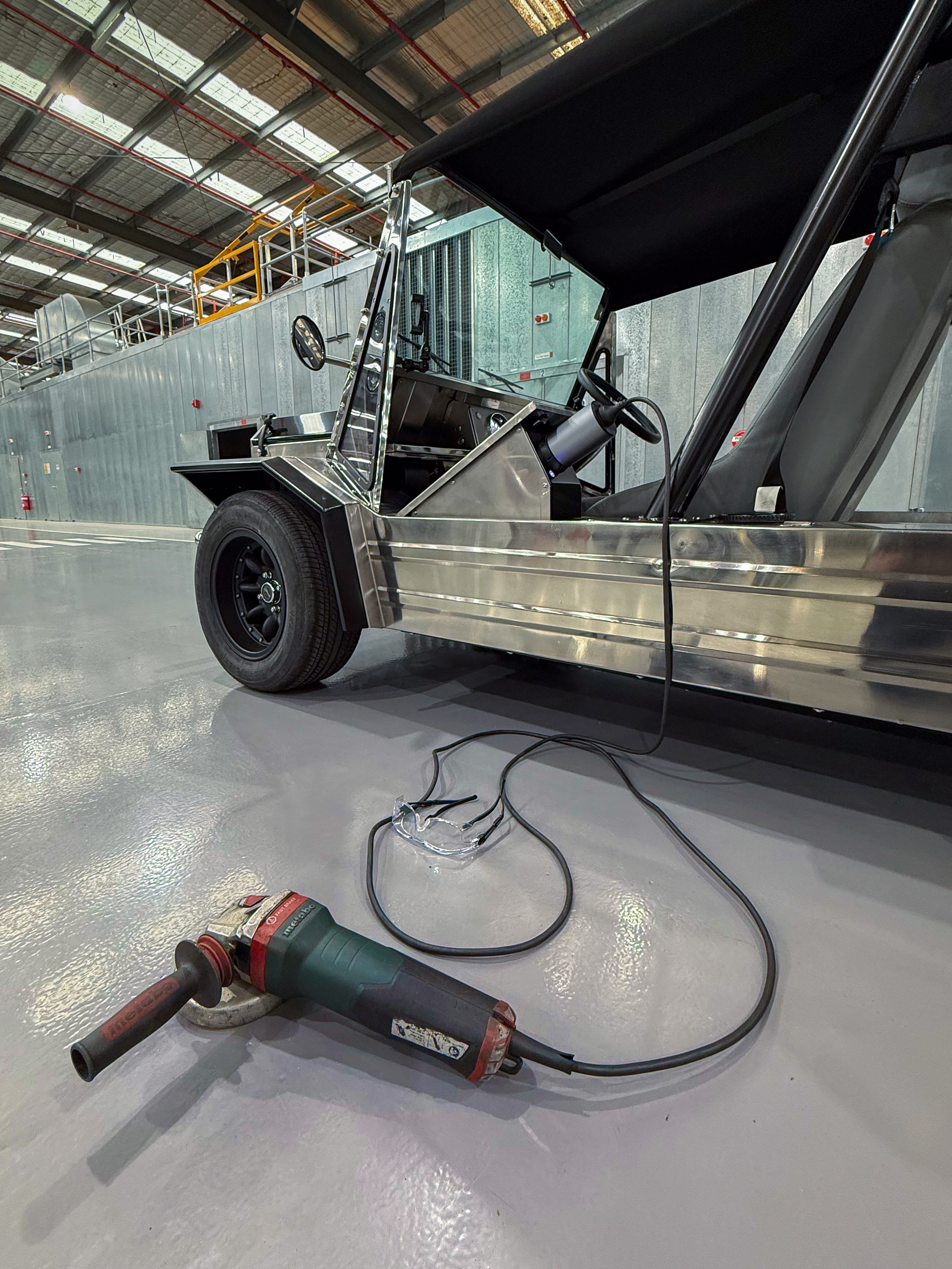 Rear three-quarter workshop view with an angle grinder resting on the floor in the foreground, the stainless body and canvas top visible behind it.