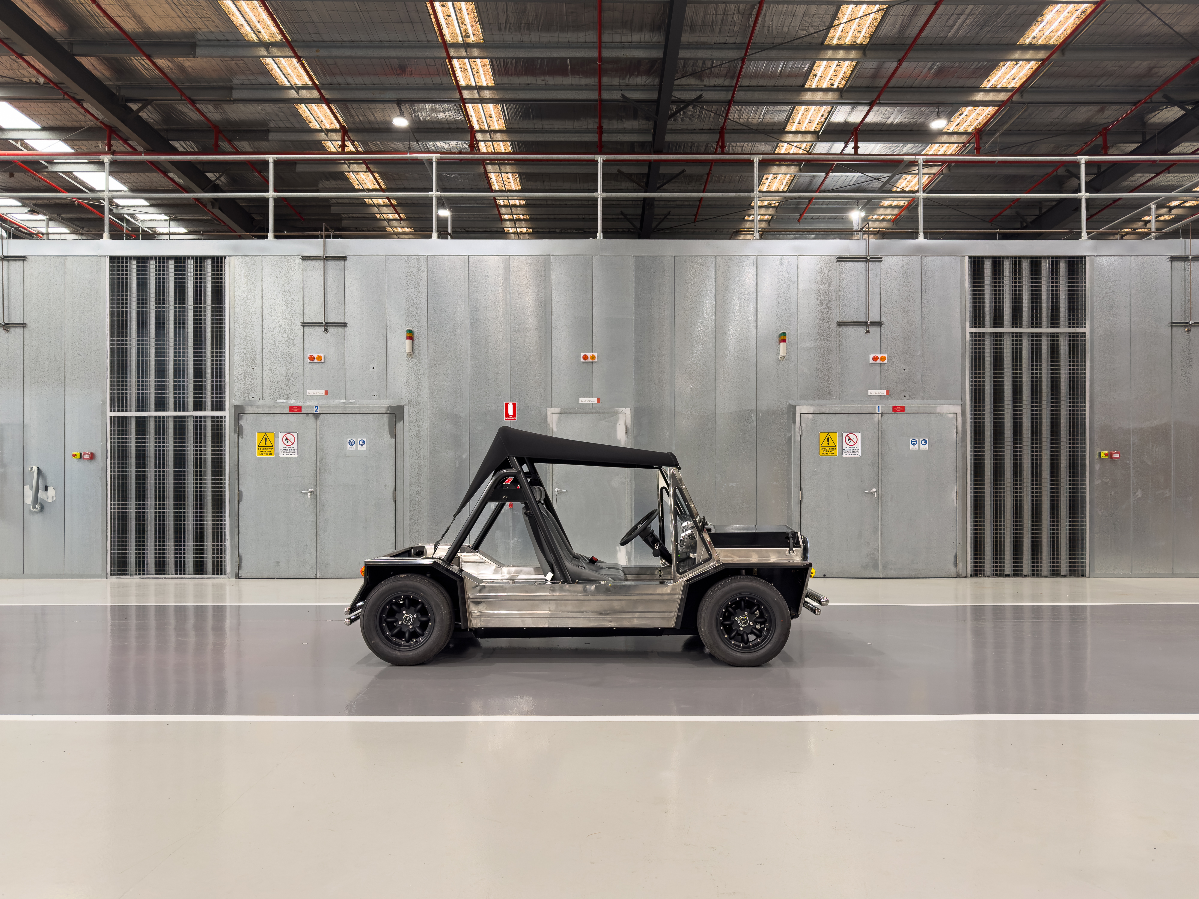 Full side profile of the stainless Moke centred on the factory floor against galvanised electrical cabinet walls, fluorescent lighting overhead.