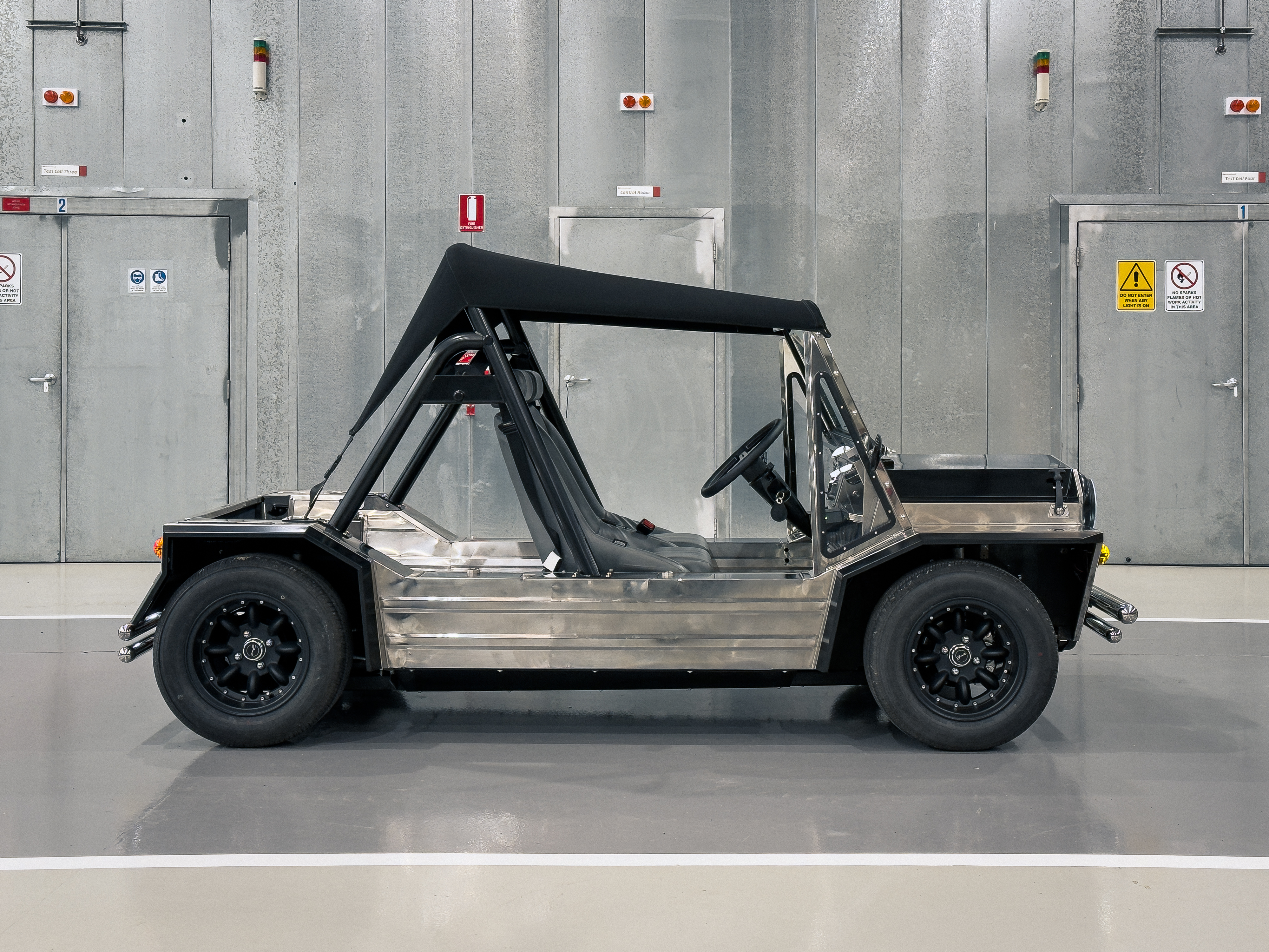Closer side-profile shot of the completed Moke with canvas top up, showing the polished body and black alloy wheels against the industrial workshop backdrop.