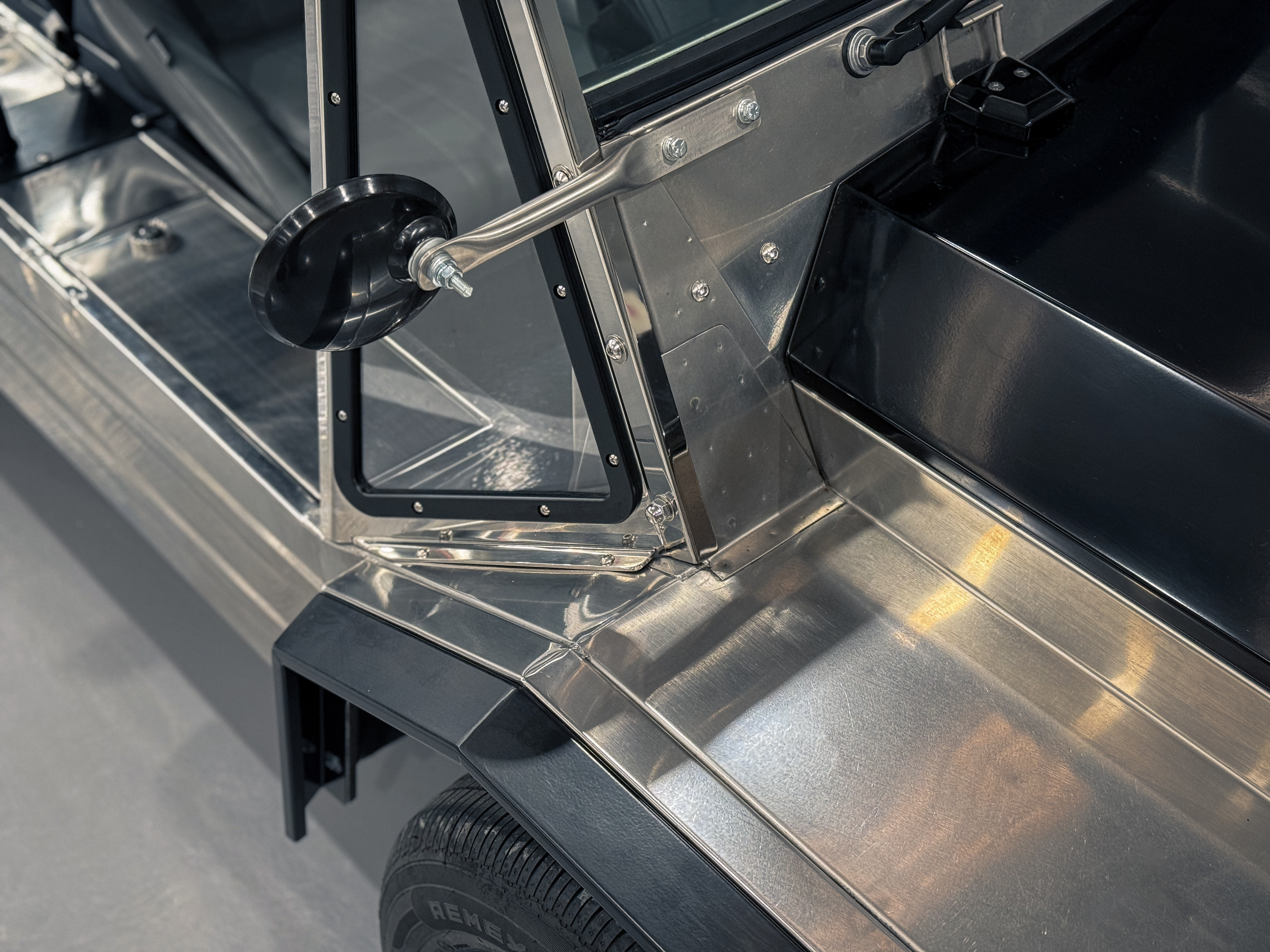 Workshop detail of the windscreen frame, side mirror, and polished stainless door sill and body panel, showing the mirror-finish of the completed body work.